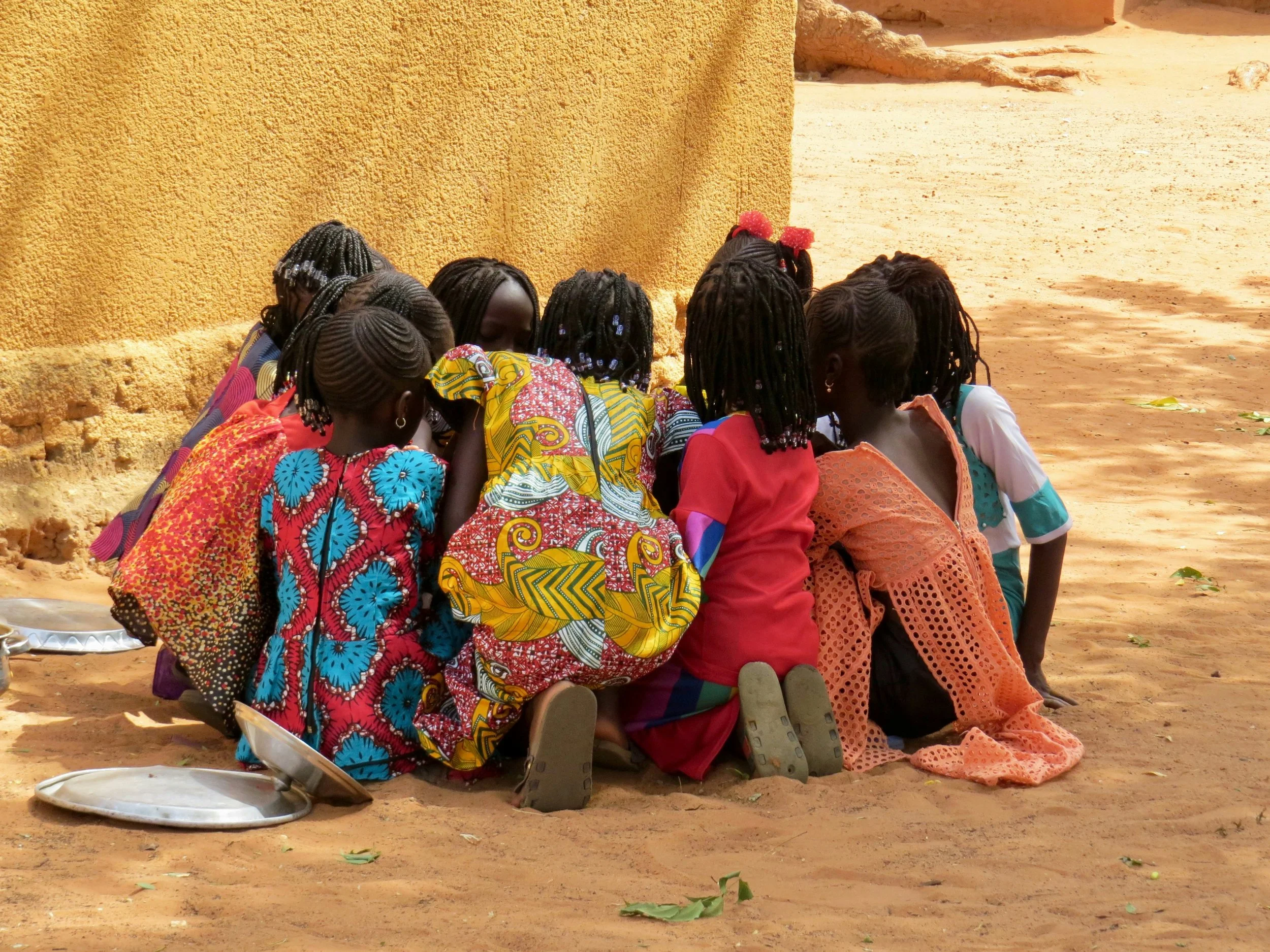 A group of children wearing colorful clothing sitting closely together on the ground near a yellow wall.