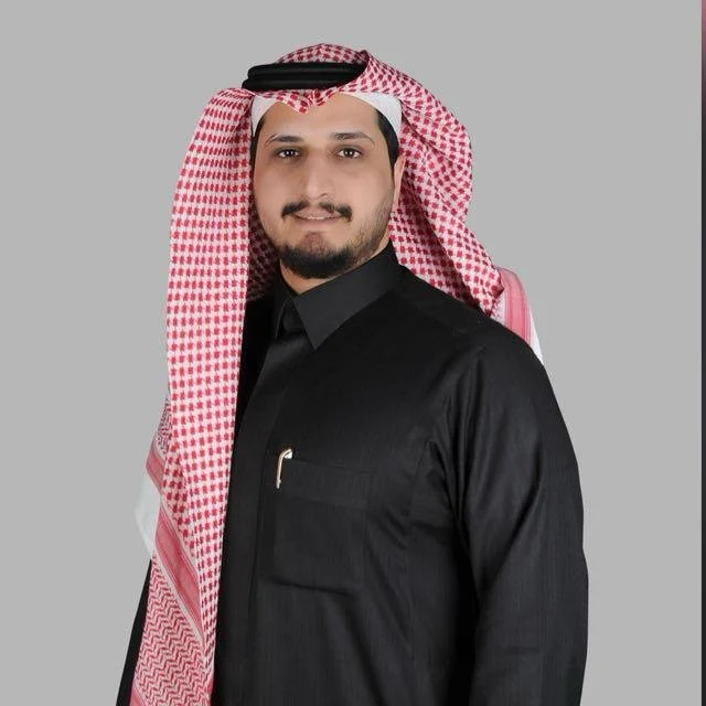 A man wearing a black traditional outfit with a red and white checkered headscarf and a black headband, posing against a plain gray background.