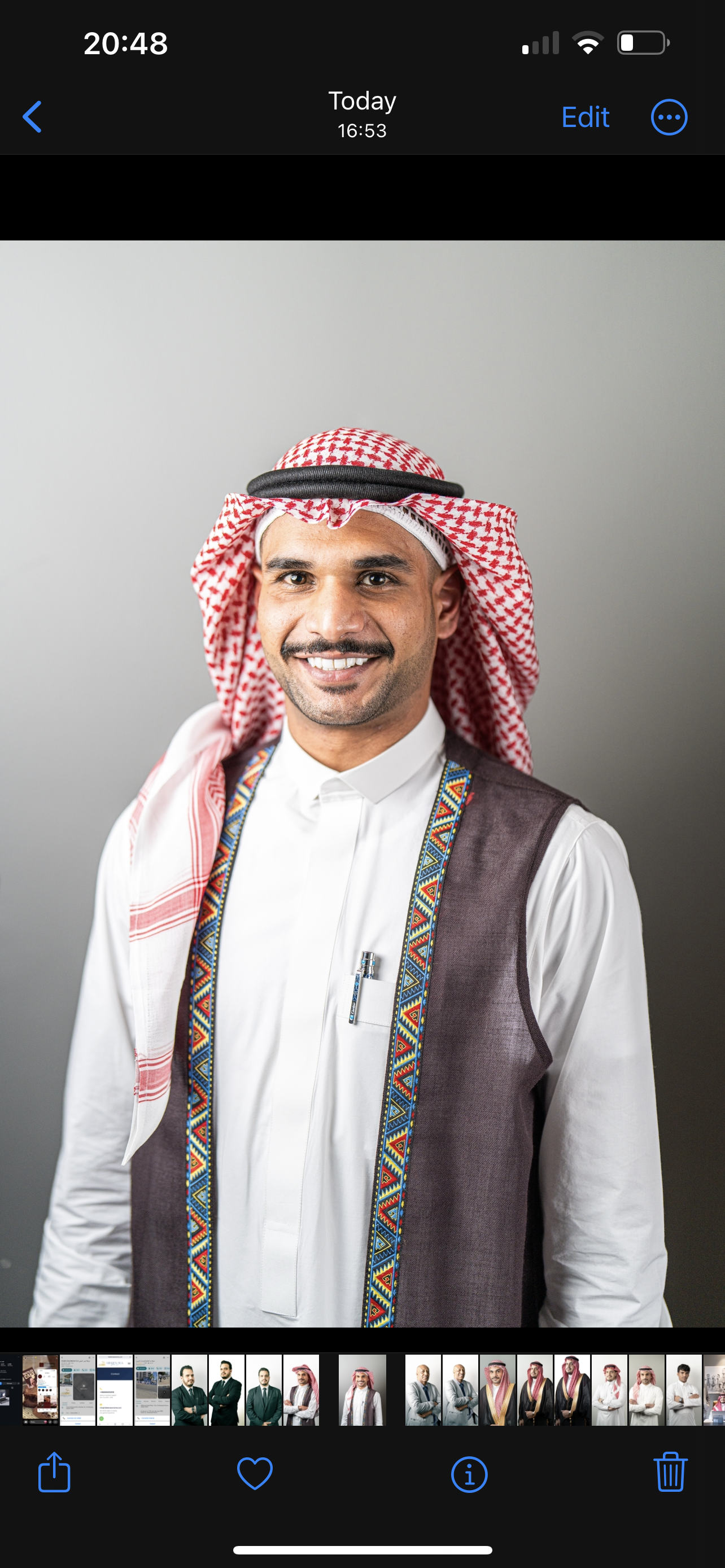 A man wearing traditional Middle Eastern attire, including a red and white checkered keffiyeh and a white thobe, with a decorative vest and a colorful patterned sash. He is smiling and standing against a neutral gray background.