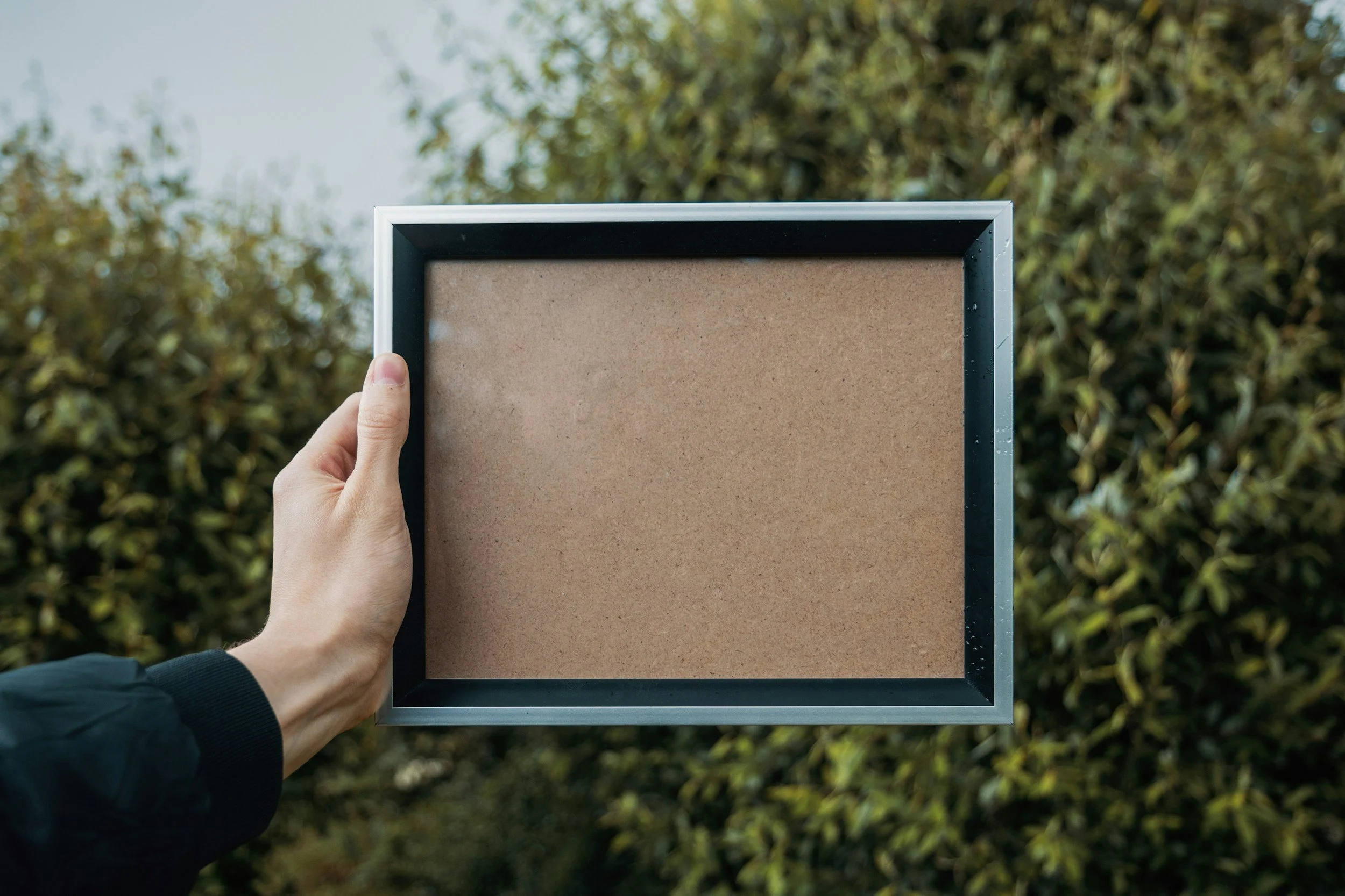 Person holding an empty black-framed corkboard outdoors with trees in the background.