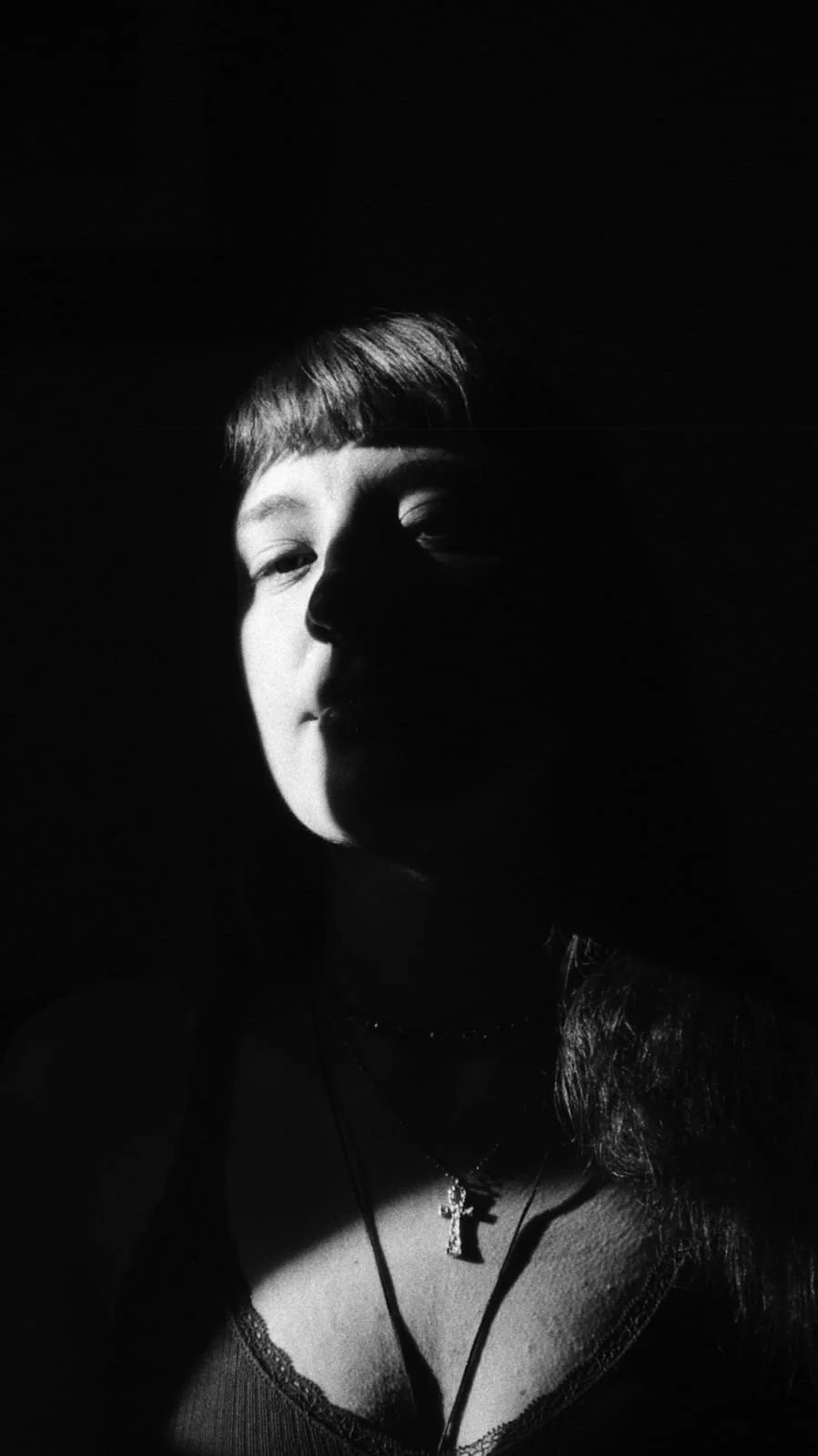 A black and white photo of a girl with half her face illuminated by light and half in shadow, wearing a necklace with a cross pendant.