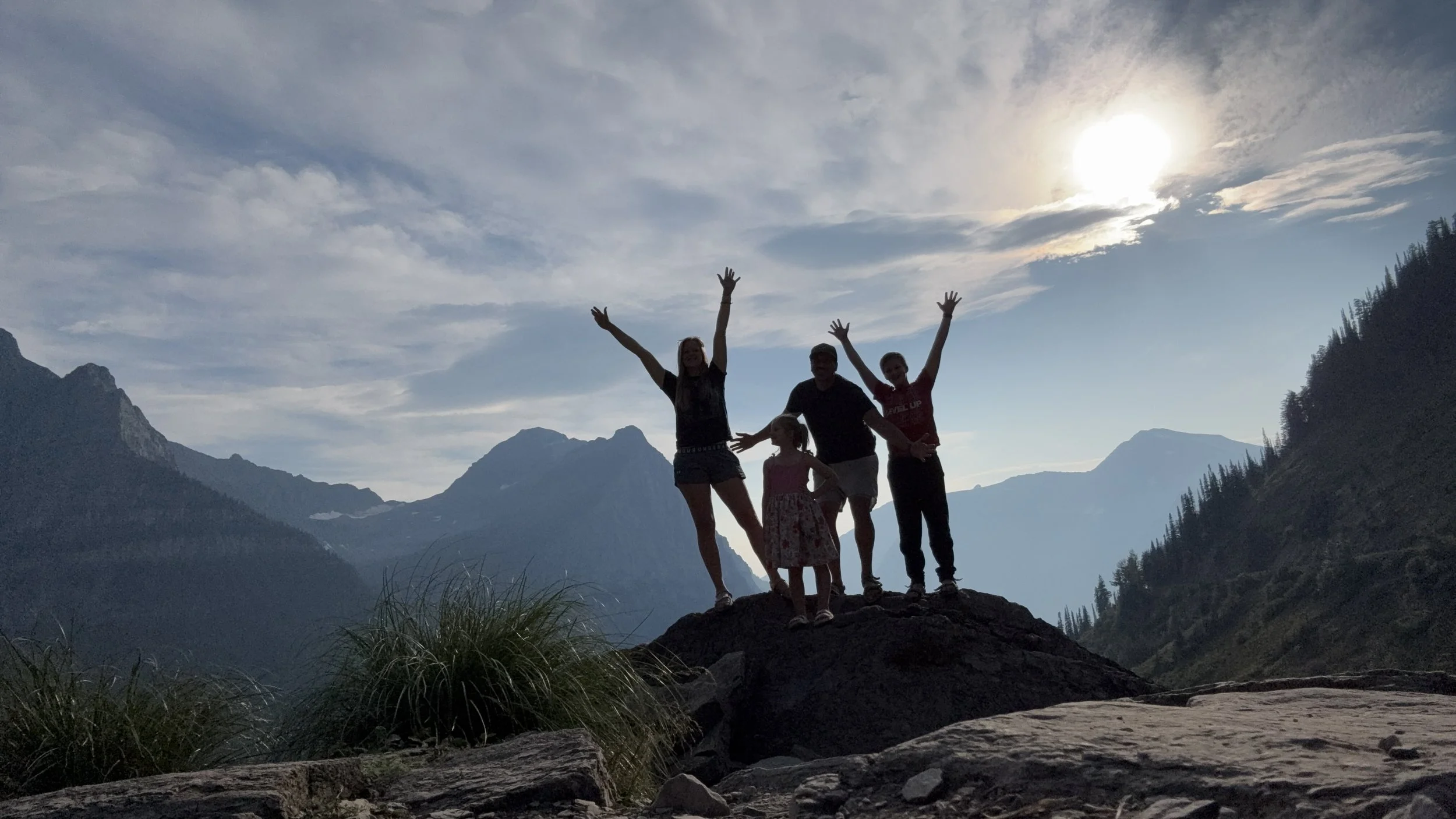 Silhouetted group of four people, including two children, standing on a rock with mountains in the background under a partly cloudy sky during sunset or sunrise.