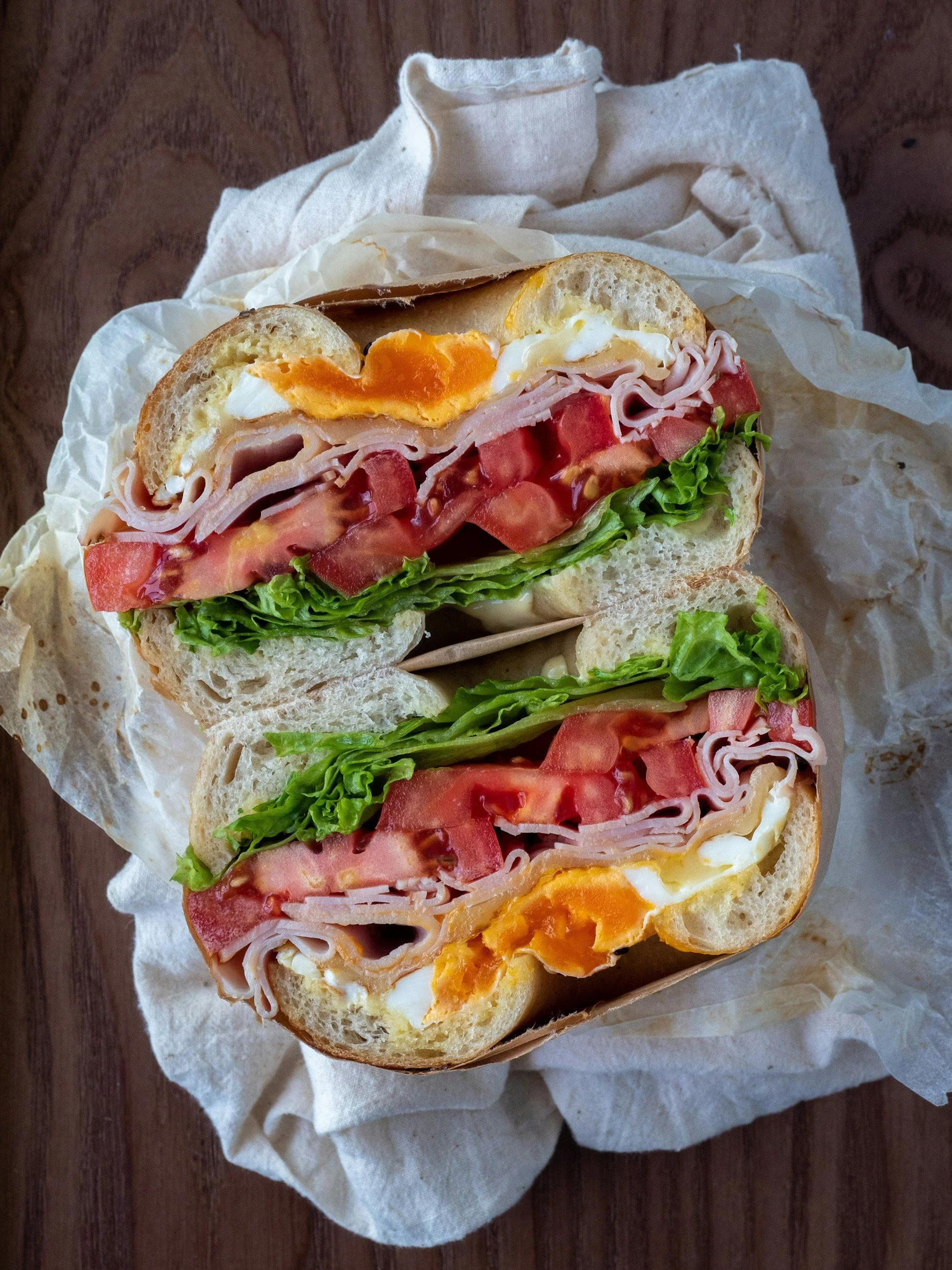 A breakfast sandwich cut in half, showing eggs, ham, tomatoes, lettuce, and bread.