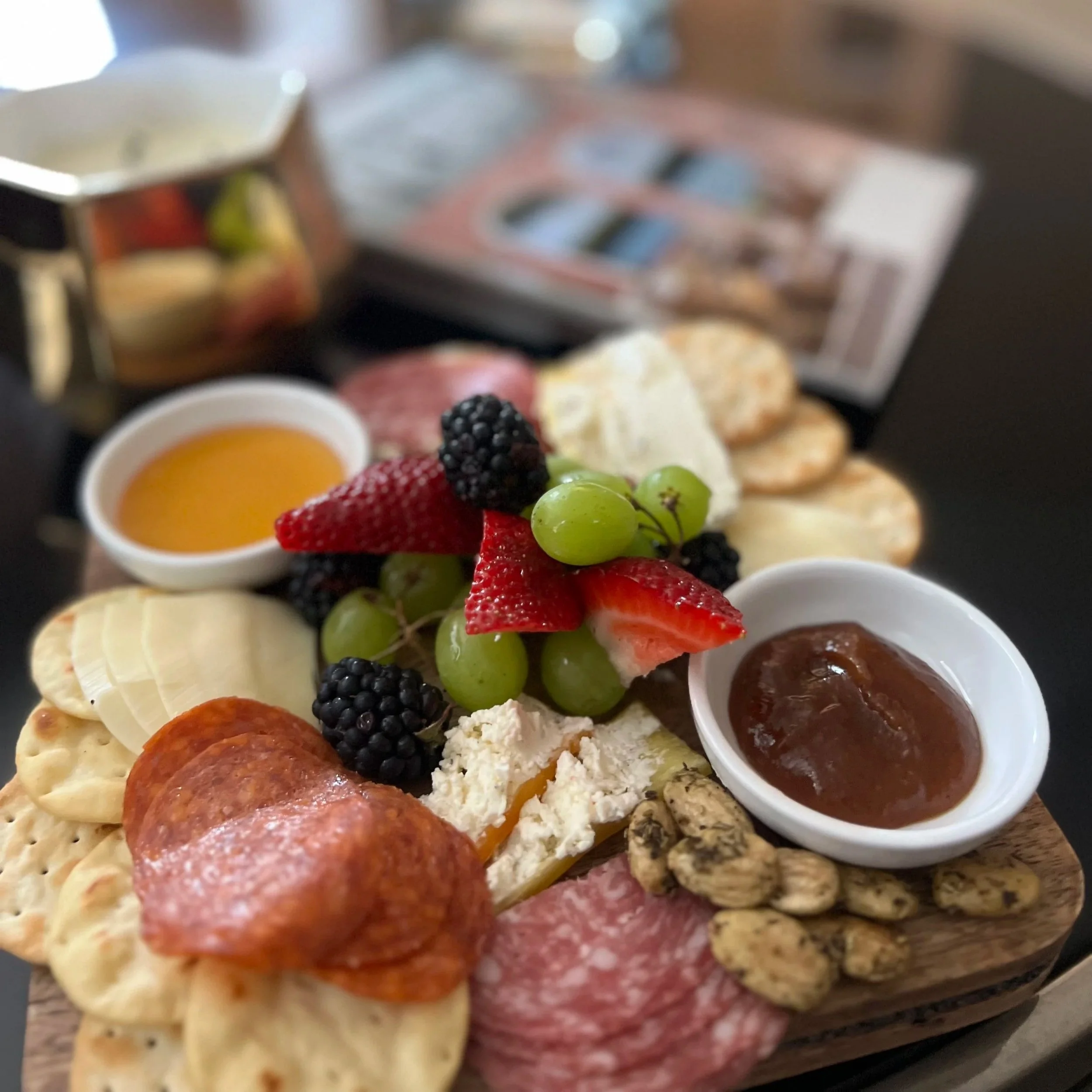 A wooden serving board with assorted breakfast items including fresh berries, sliced bananas, cheese, meats, crackers, two small bowls of jam and honey, and some peanut or nut clusters.