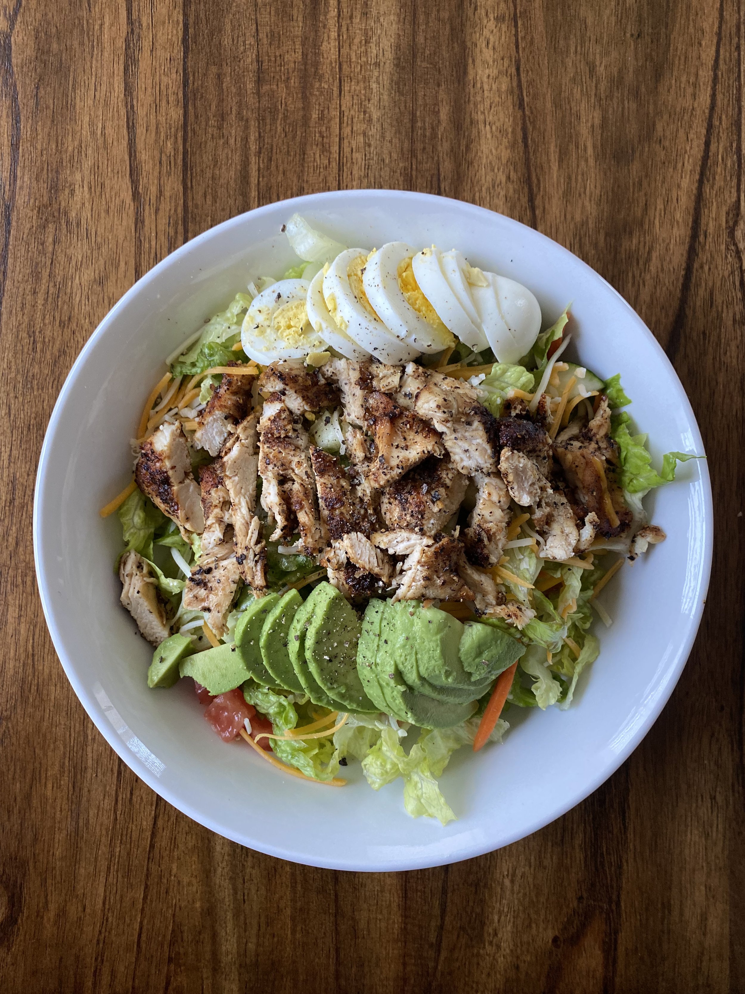 A bowl of salad with grilled chicken, sliced avocado, chopped lettuce, shredded cheese, and sliced boiled egg on top, served on a wooden table.