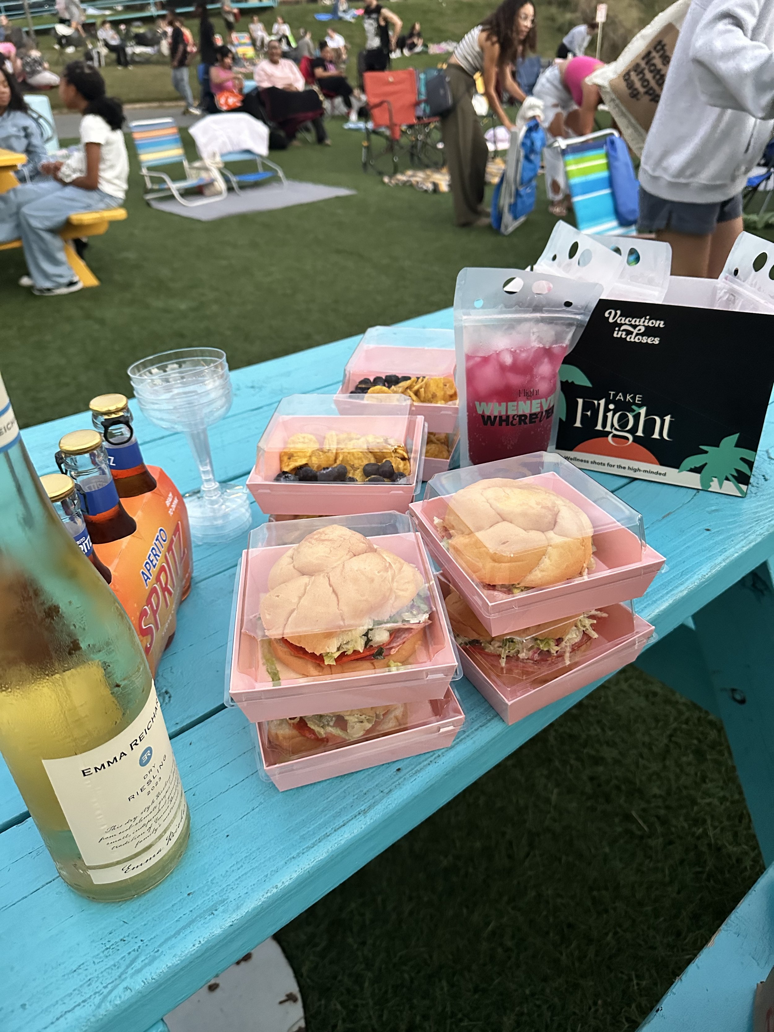 A bright blue outdoor picnic table set with wrapped sandwiches, snacks, bottled drinks, and a glass, with a crowd of people sitting and walking in a park in the background.