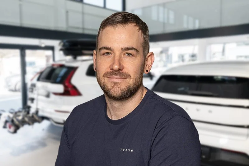 Porträt eines lächelnden Mannes mit kurzen braunen Haaren, Bart und einem schwarzen Zip-Up-Hoodie vor einem hellen Hintergrund.