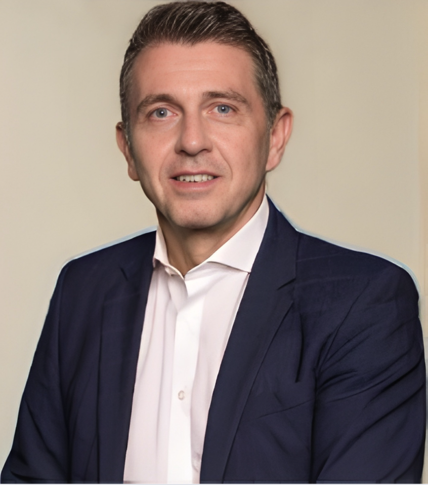 Ein Mann trägt einen dunklen Anzug und ein weißes Hemd, lächelt leicht, vor hellem Hintergrund fotografiert.