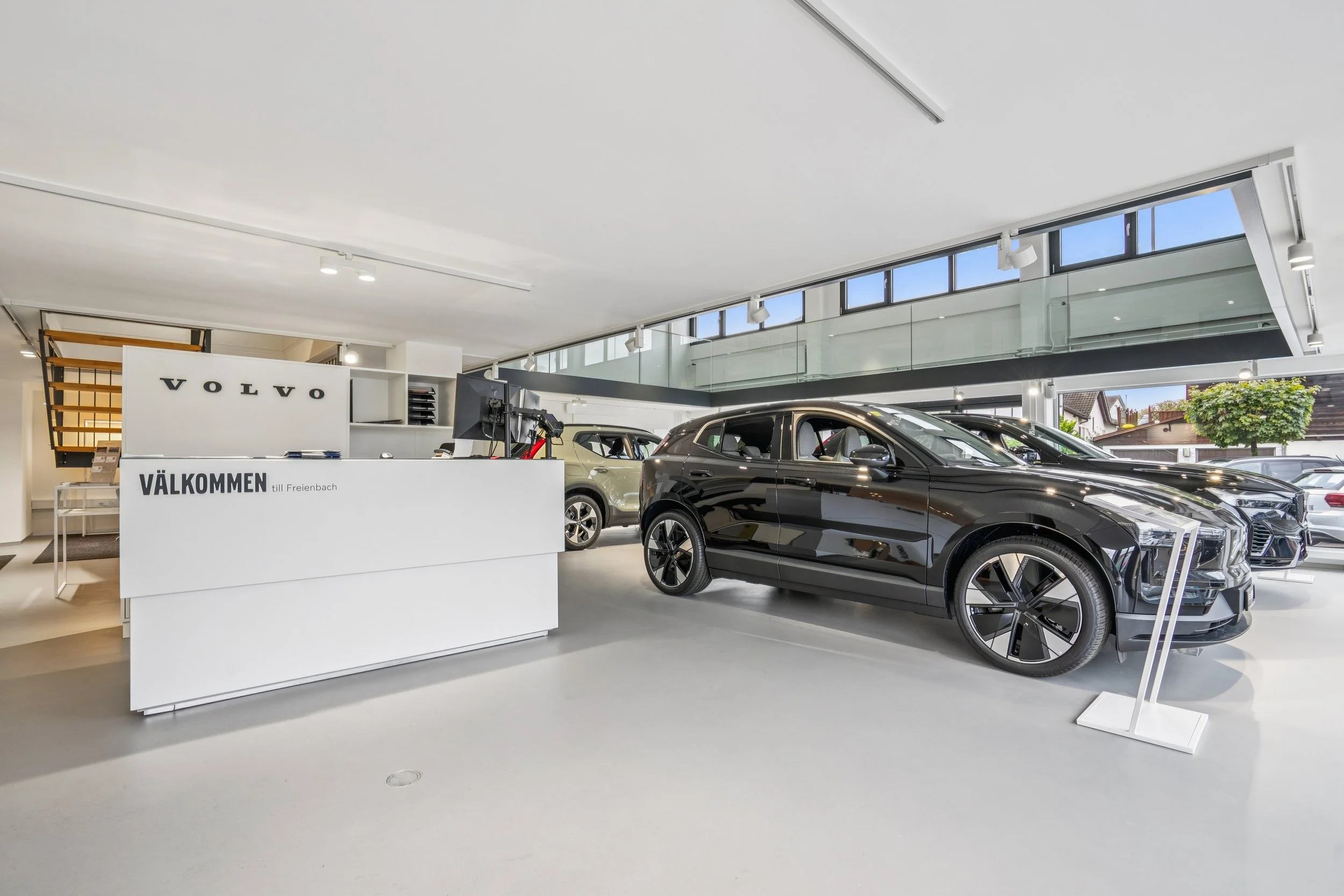 Innenansicht eines Autohauses mit mehreren schwarzen und weißen Autos, einem Empfangstresen mit der Beschriftung 'VÄLKOMMEN' und einem Volvo-Logo im Hintergrund.