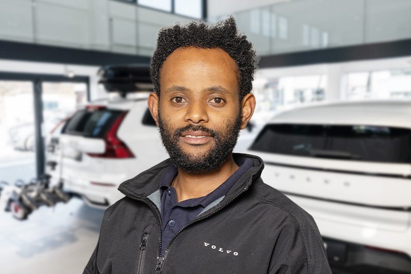 Porträt eines lachenden Mannes mit dunklem Haar, Bart und schwarz Kleidung vor hellem Hintergrund.