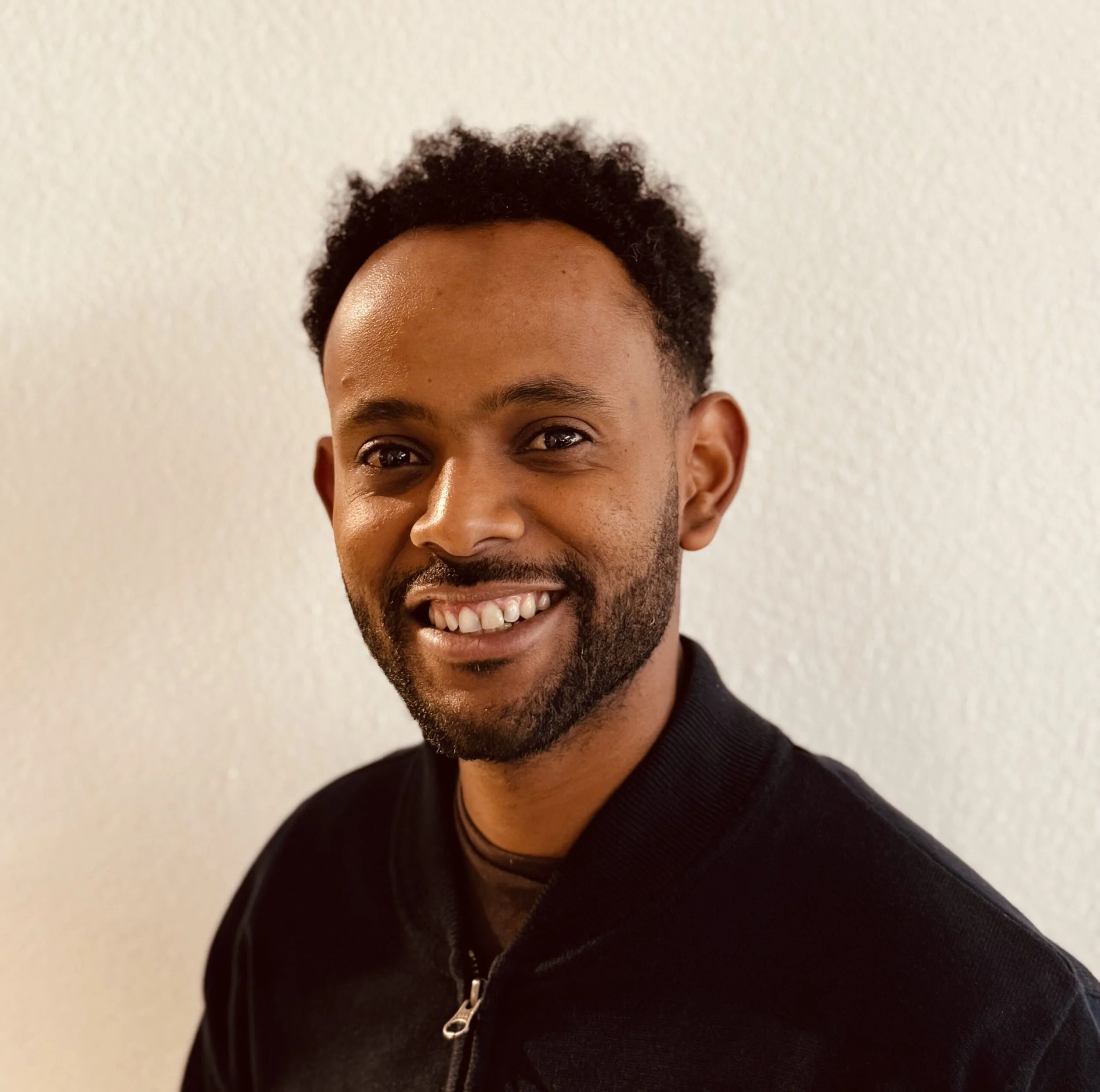 Porträt eines lachenden Mannes mit dunklem Haar, Bart und schwarz Kleidung vor hellem Hintergrund.