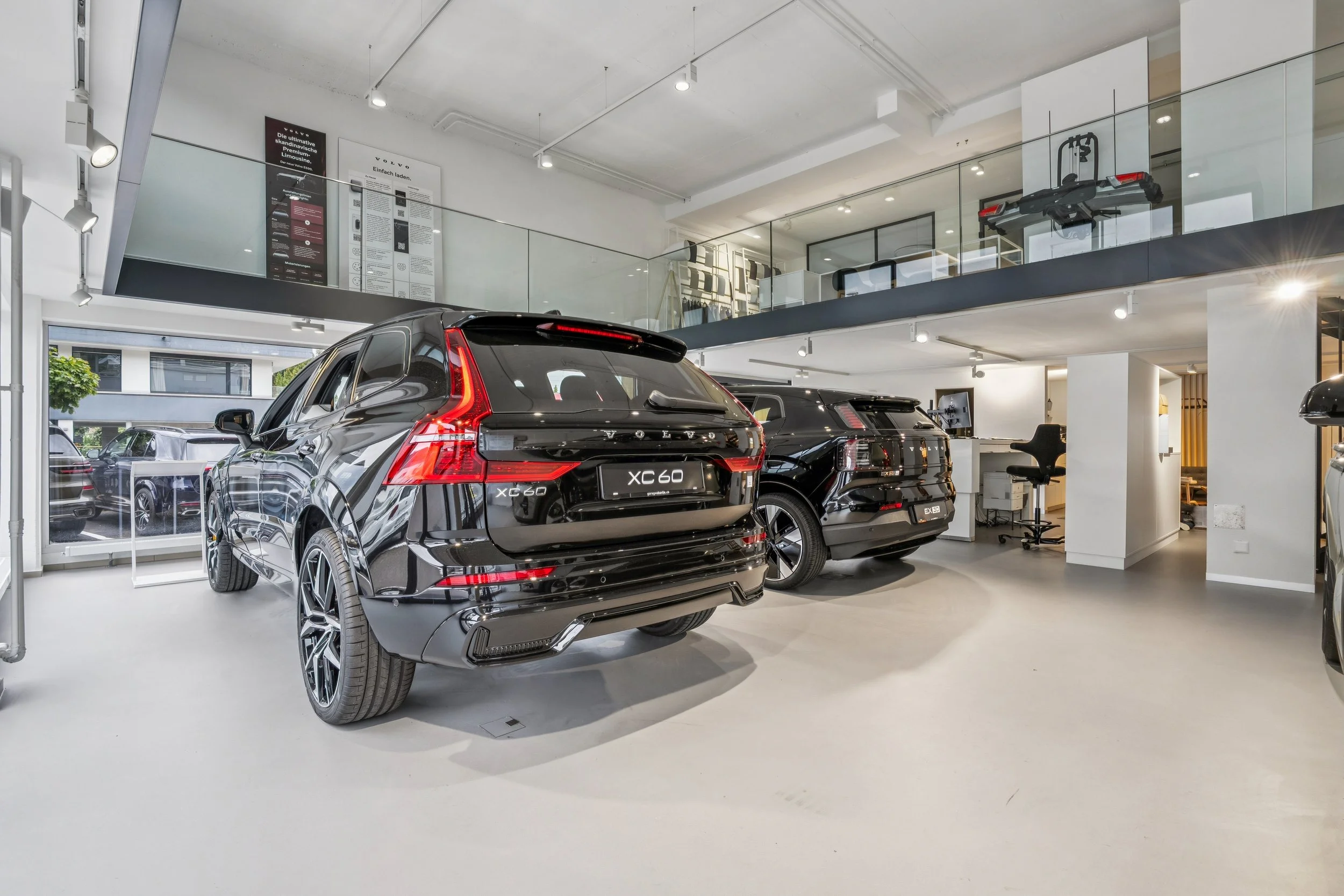 Zwei schwarze Volvo XC60 SUVs in einem modernen Autohandelstore, mit einem Bürobereich im Hintergrund und Fenstern, die Tageslicht hereinlassen.