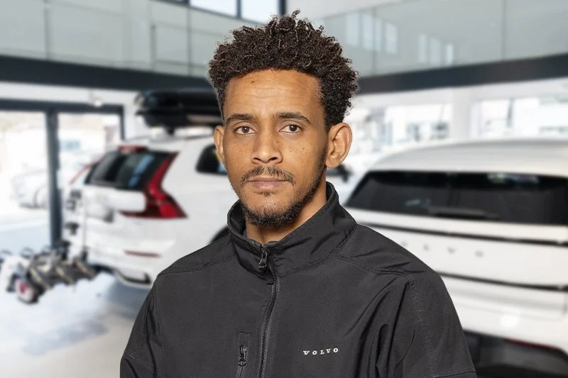 Portrait eines lächelnden Mannes mit dunklem Teint, lockigem Haar und kurzem Bart, trägt eine schwarze Jacke vor einer weißen Wand.