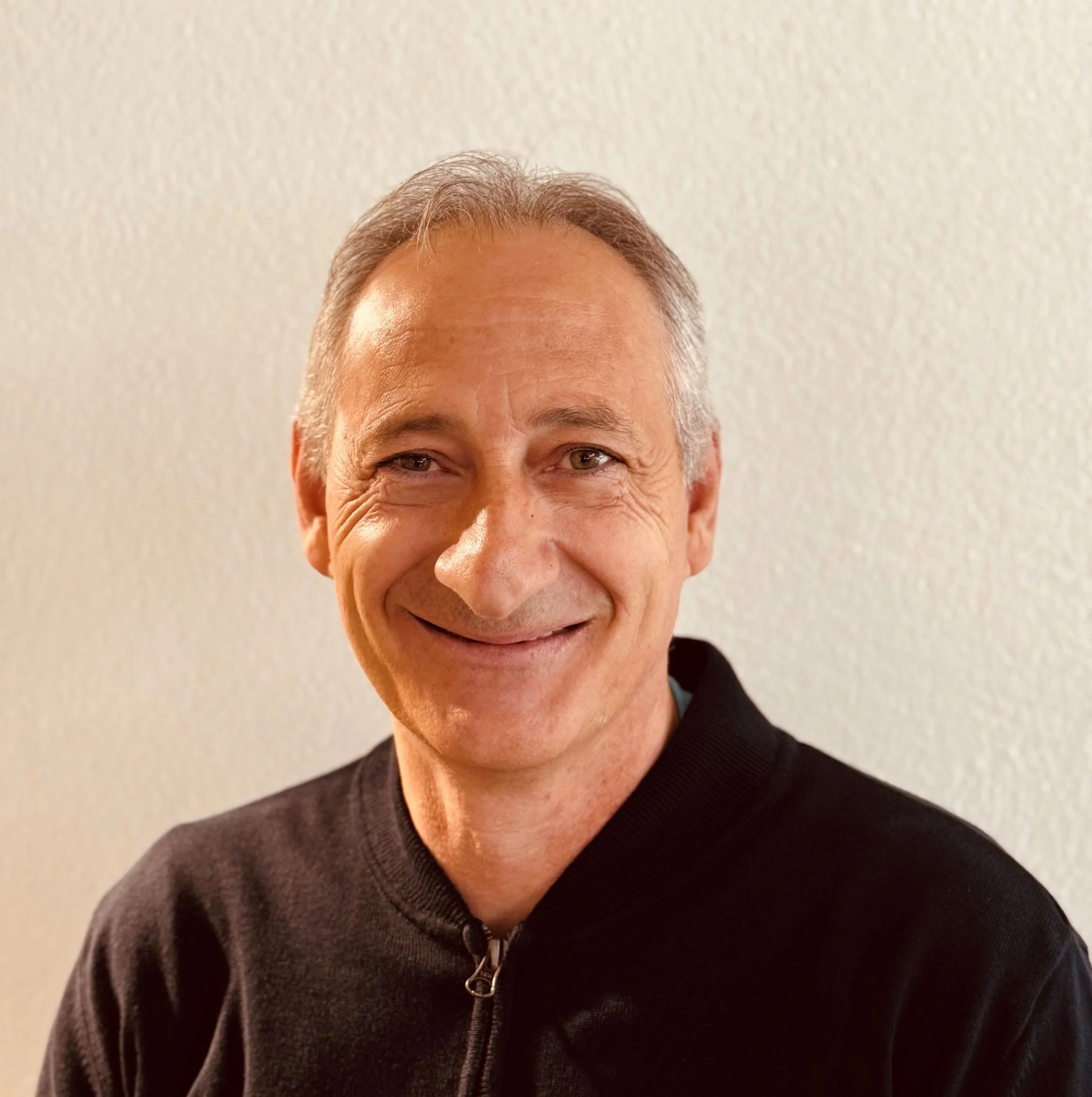 Ein Mann mit grauen Haaren trägt einen schwarzen Pullover und lächelt.