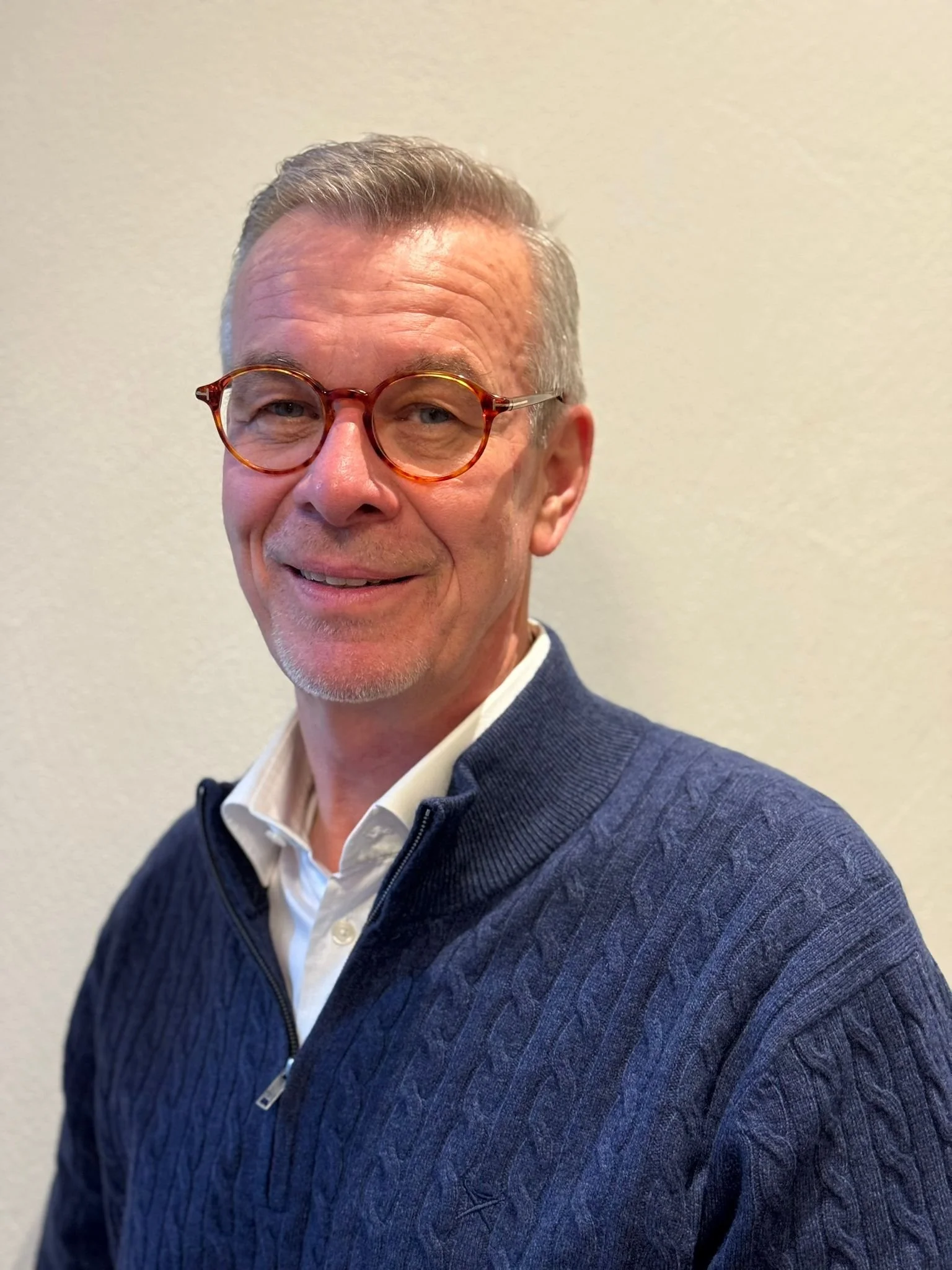 Ein Mann mit kurzen, grauen Haaren, Brille mit runden Gestellen, dunklem Pullover und weißem Hemd steht vor einem weißen Hintergrund, lächelt.