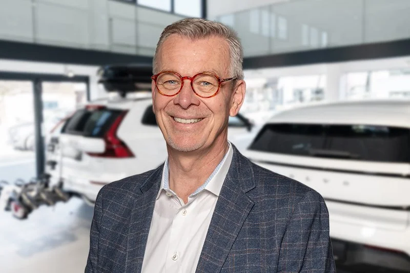 Ein Mann mit kurzen, grauen Haaren, Brille mit runden Gestellen, dunklem Pullover und weißem Hemd steht vor einem weißen Hintergrund, lächelt.