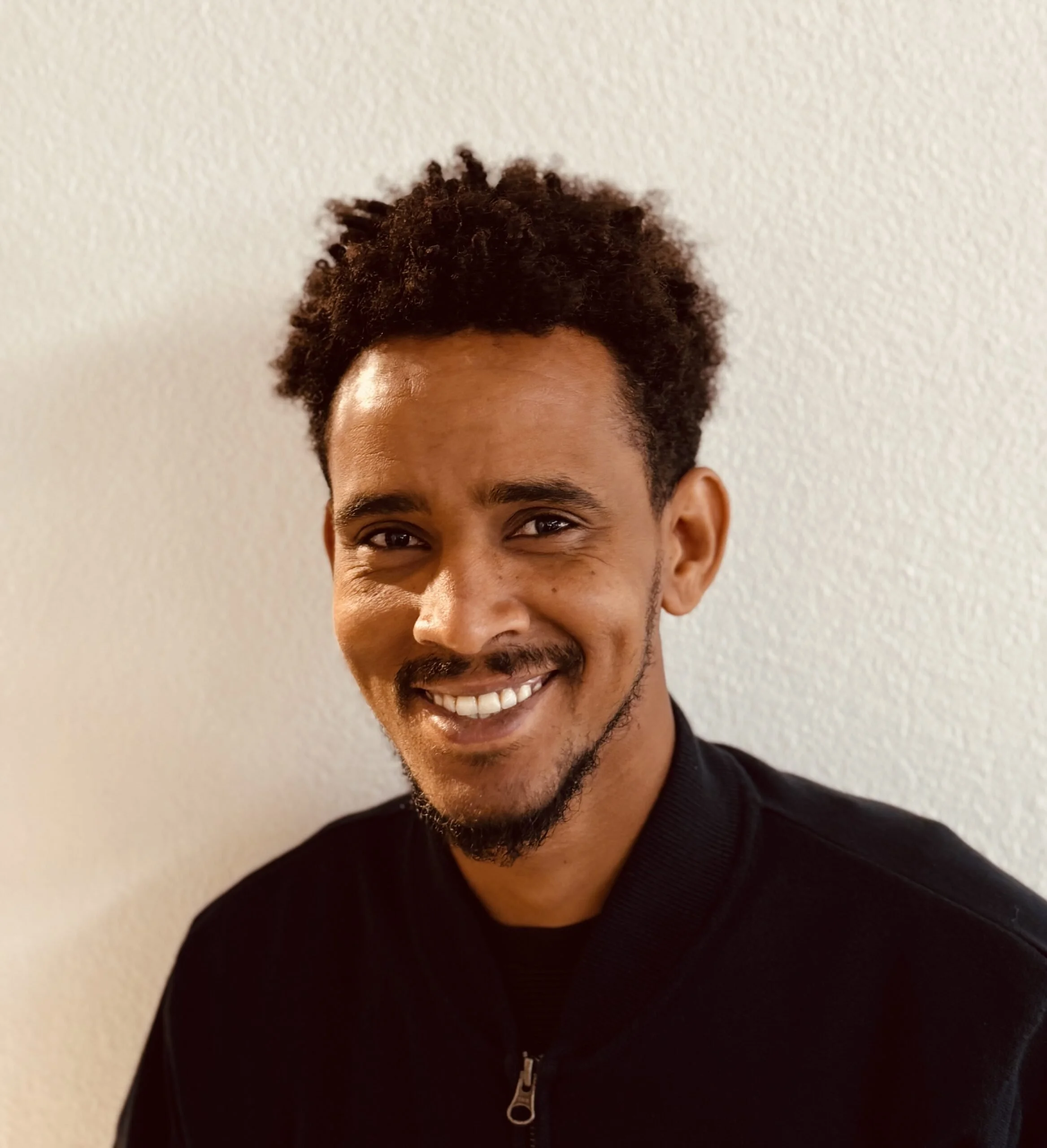 Portrait eines lächelnden Mannes mit dunklem Teint, lockigem Haar und kurzem Bart, trägt eine schwarze Jacke vor einer weißen Wand.