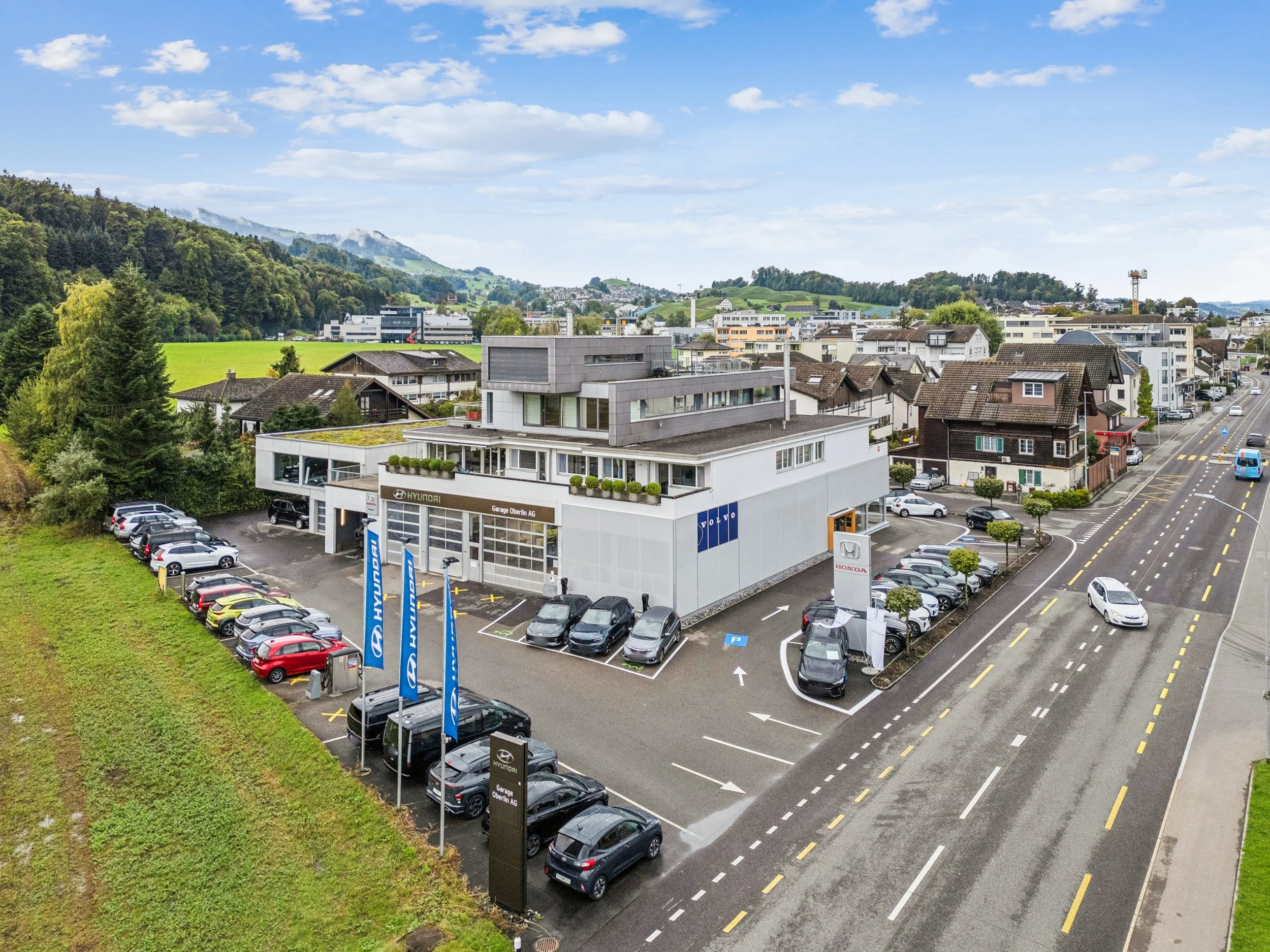 Autohaus mit Hyundai, Honda und Volvo Logos, Parkplätze, Straße, Wohnhausanwesen in der Nähe, Bergen im Hintergrund, bewölkter Himmel.