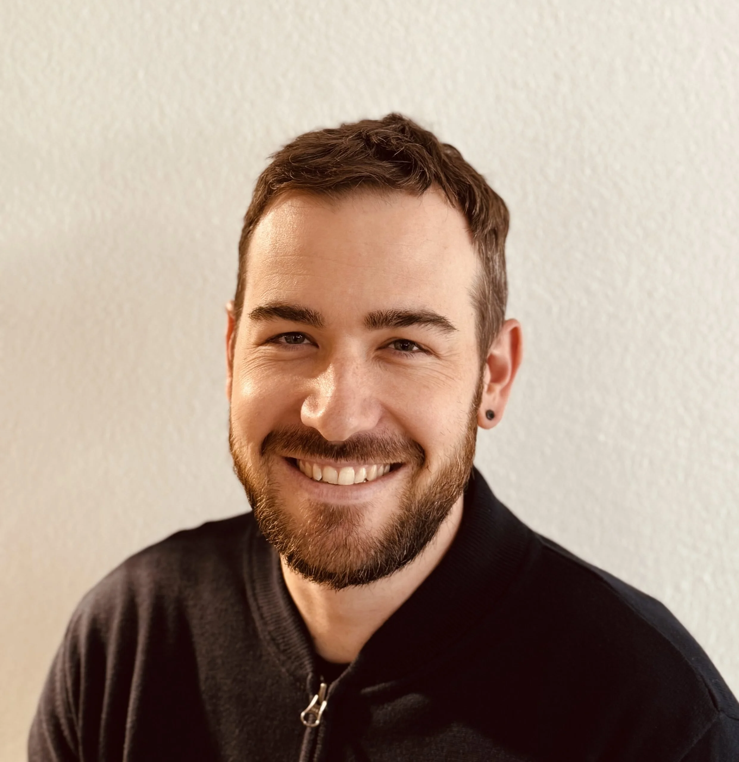 Porträt eines lächelnden Mannes mit kurzen braunen Haaren, Bart und einem schwarzen Zip-Up-Hoodie vor einem hellen Hintergrund.