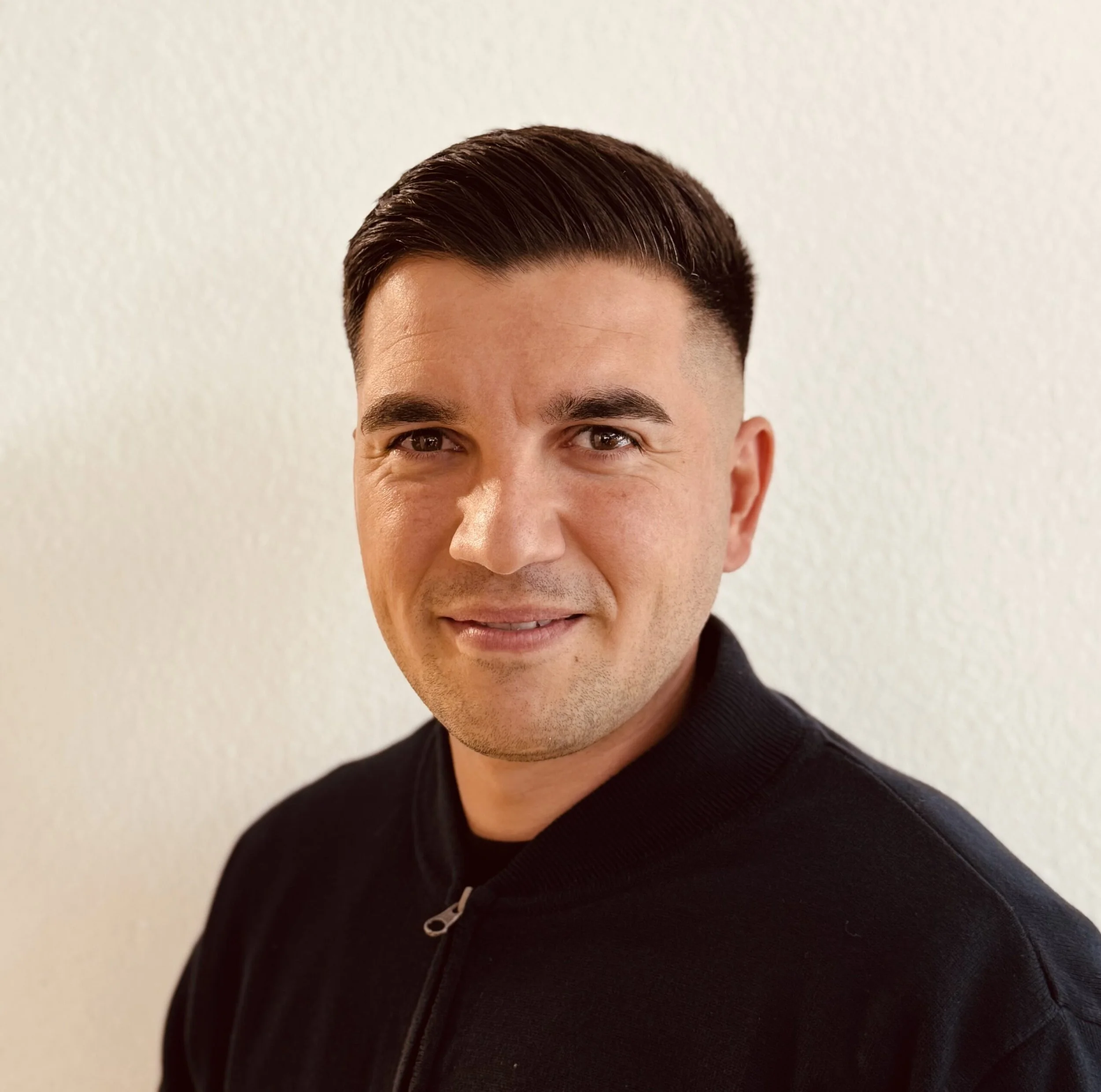 Porträt eines Mannes mit dunklen Haaren vor weißer Wand, trägt eine schwarze Jacke.