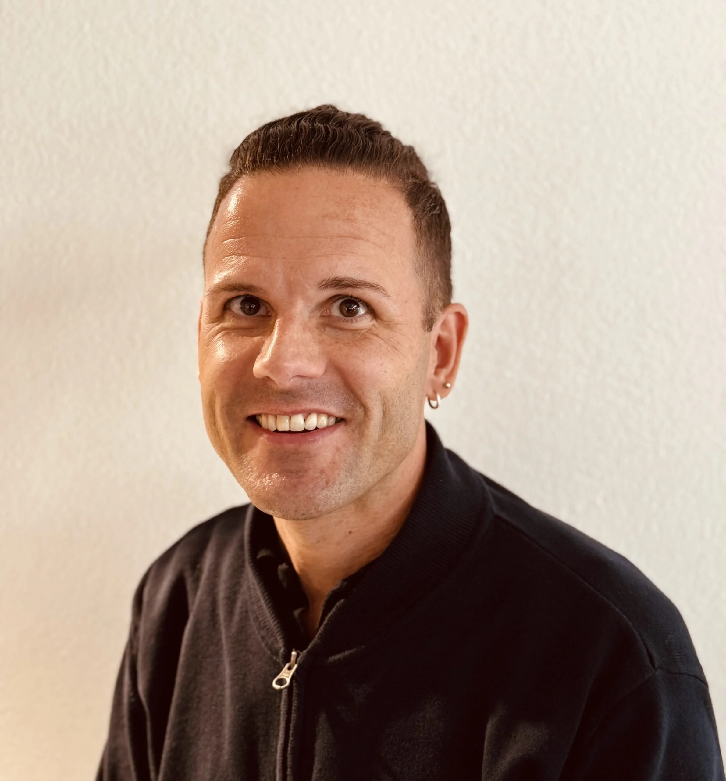 Porträt eines Mannes mit dunklem Haar, Eilend, in schwarzem Zipper, vor hellem Hintergrund.