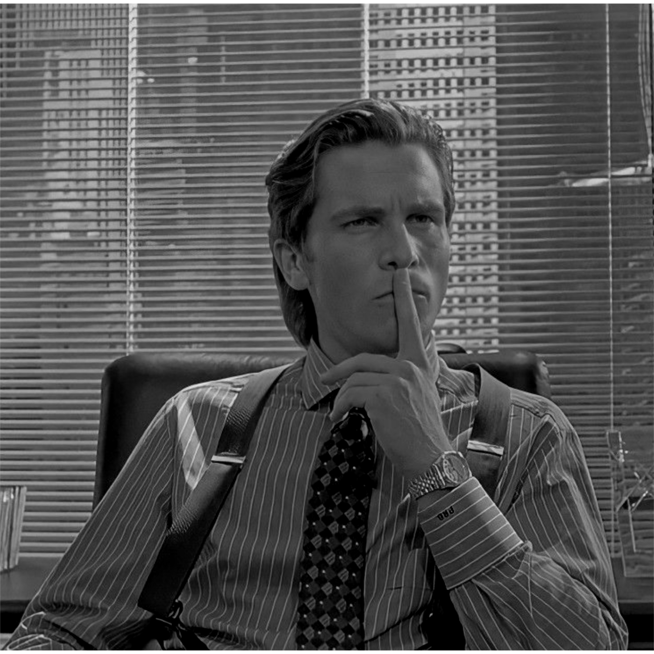 A young man in a suit and tie sitting in an office chair, holding his index finger to his lips in a 'shh' gesture. The background shows window blinds and city buildings.