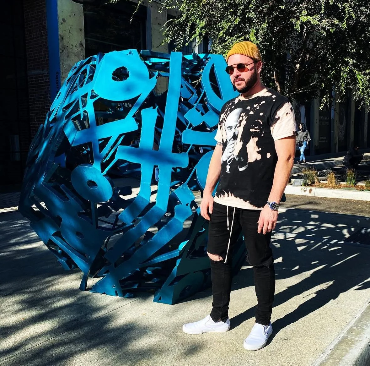 A man standing outdoors next to a modern blue metal sculpture, wearing a yellow beanie, sunglasses, a black and white graphic t-shirt, black ripped jeans, and white sneakers.