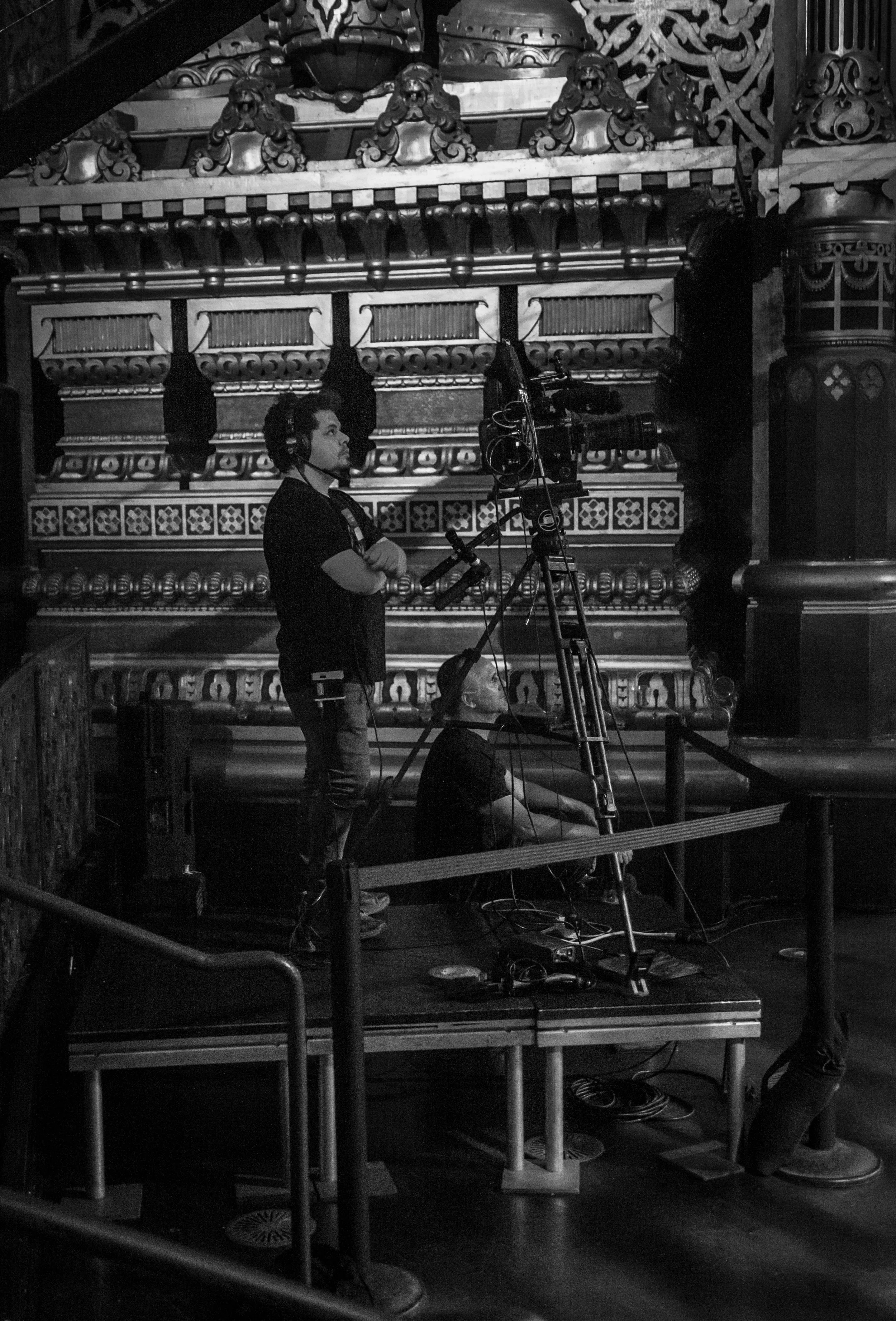 Two videographers working with a camera on a stage in a historic venue with ornate wooden decorations.