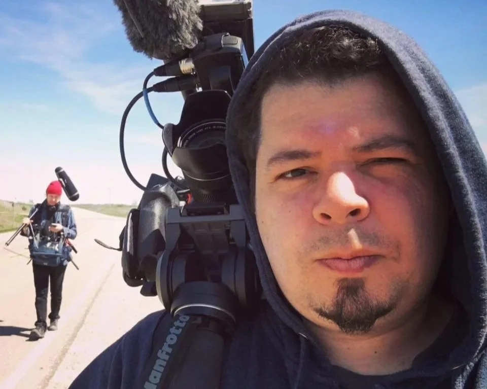 Man wearing a hoodie taking a selfie on an empty road with another person walking in the background.