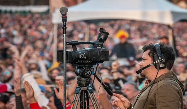 Cameraman filming at an outdoor event with a large crowd in the background.