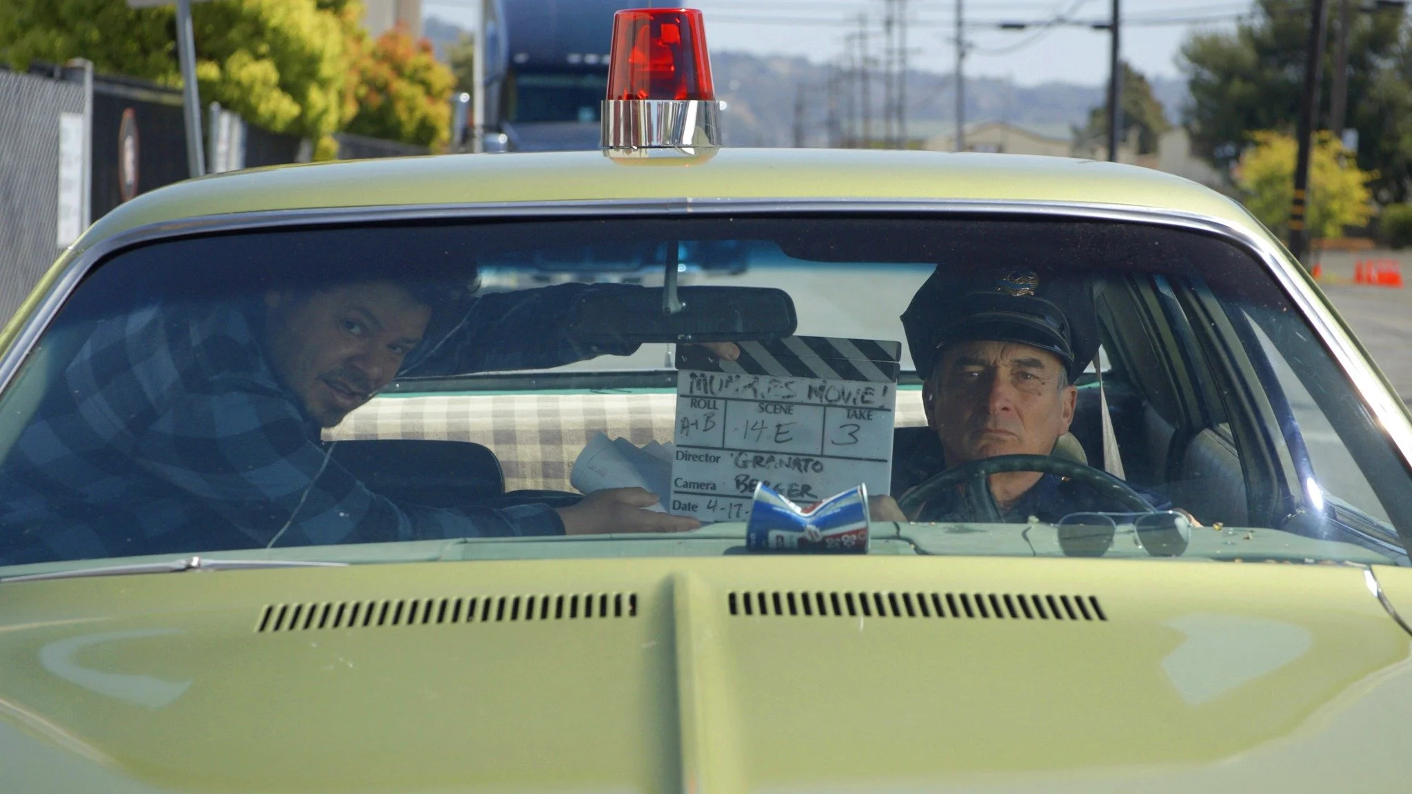 Two men sitting inside a yellow police car. One of them is the driver, wearing a police hat, looking serious. The other, in the passenger seat, is pointing a film slate towards the camera, smiling. The car has a red siren on top and sunglasses restin