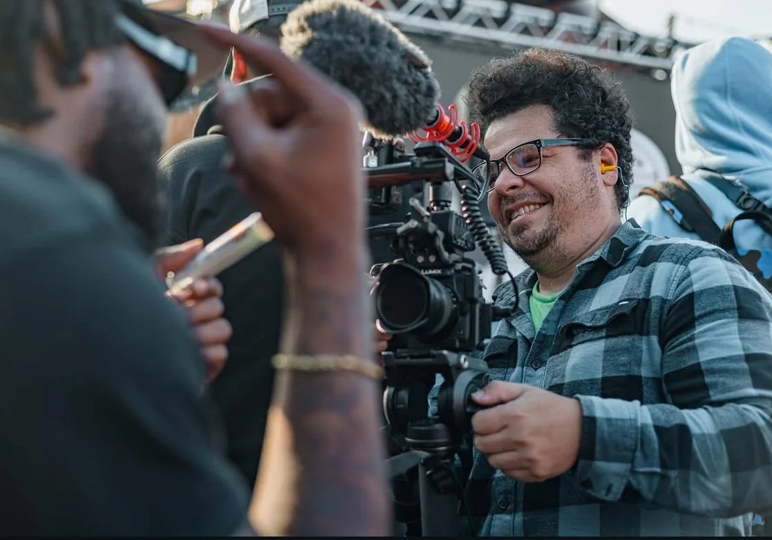 Smiling man with glasses and curly hair operating a professional camera outdoors at an event.