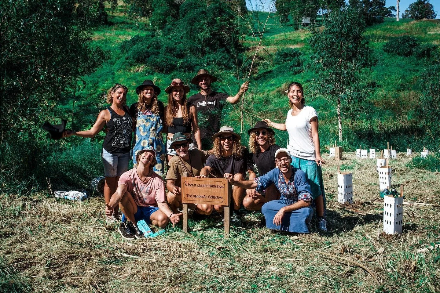 Group of people planting trees in a forested area, holding a sign that reads 'A forest planted with love. The VanderAa Collective', with small tree protectors around young trees.