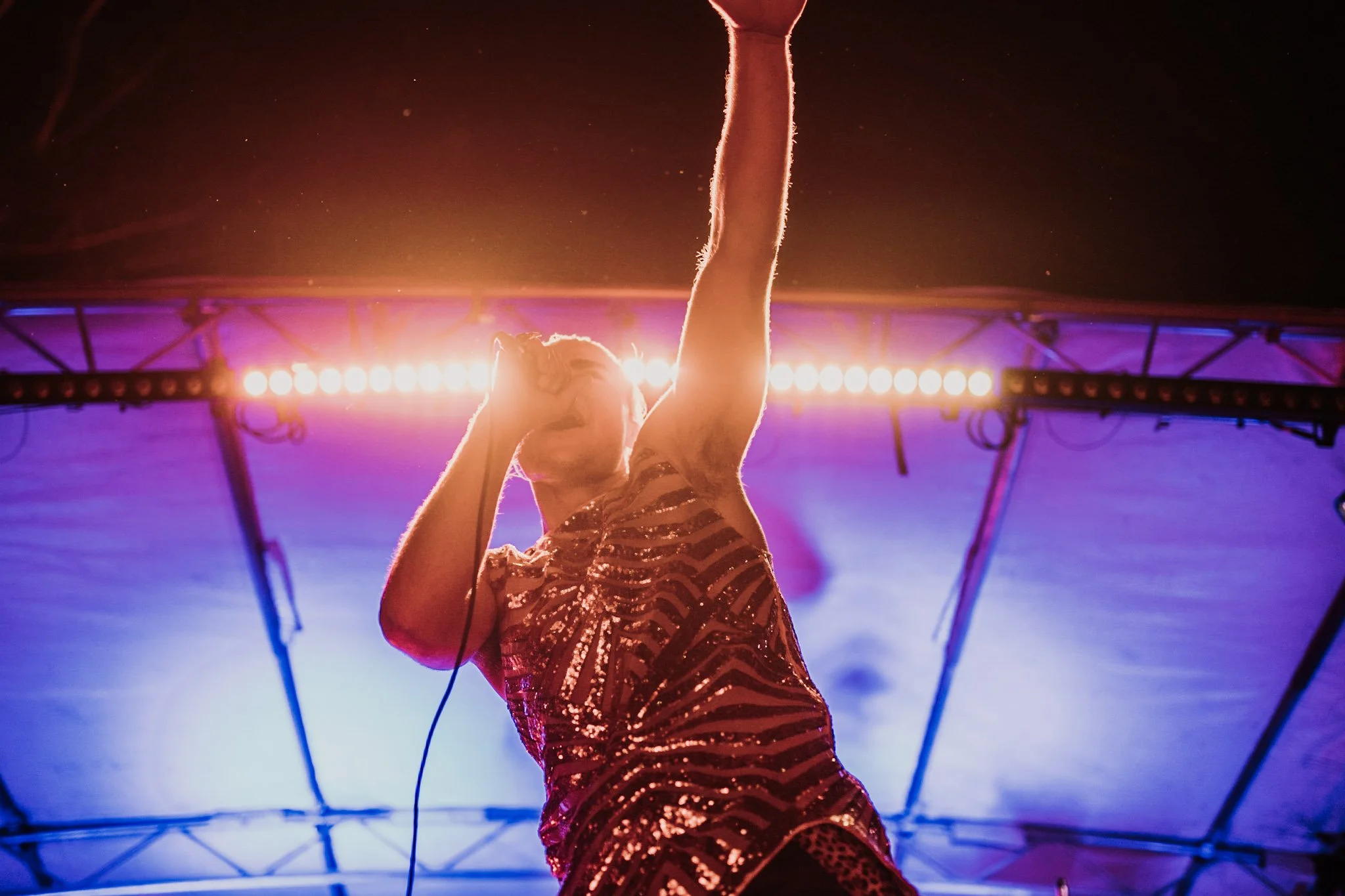 Jackson Kubian rapping into a microphone on stage, wearing a shiny, patterned outfit with a high ponytail, illuminated by colorful stage lights.