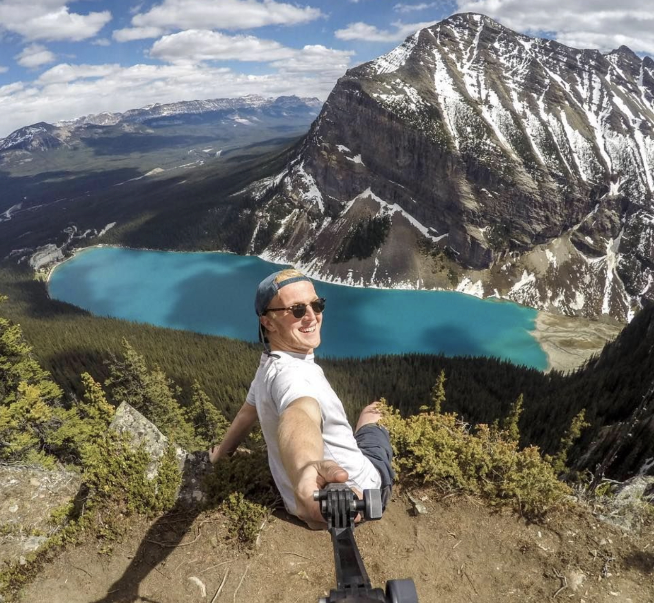 A man taking a selfie on a mountain trail, with a scenic view of a turquoise lake, forest, and snow-capped mountains in the background.