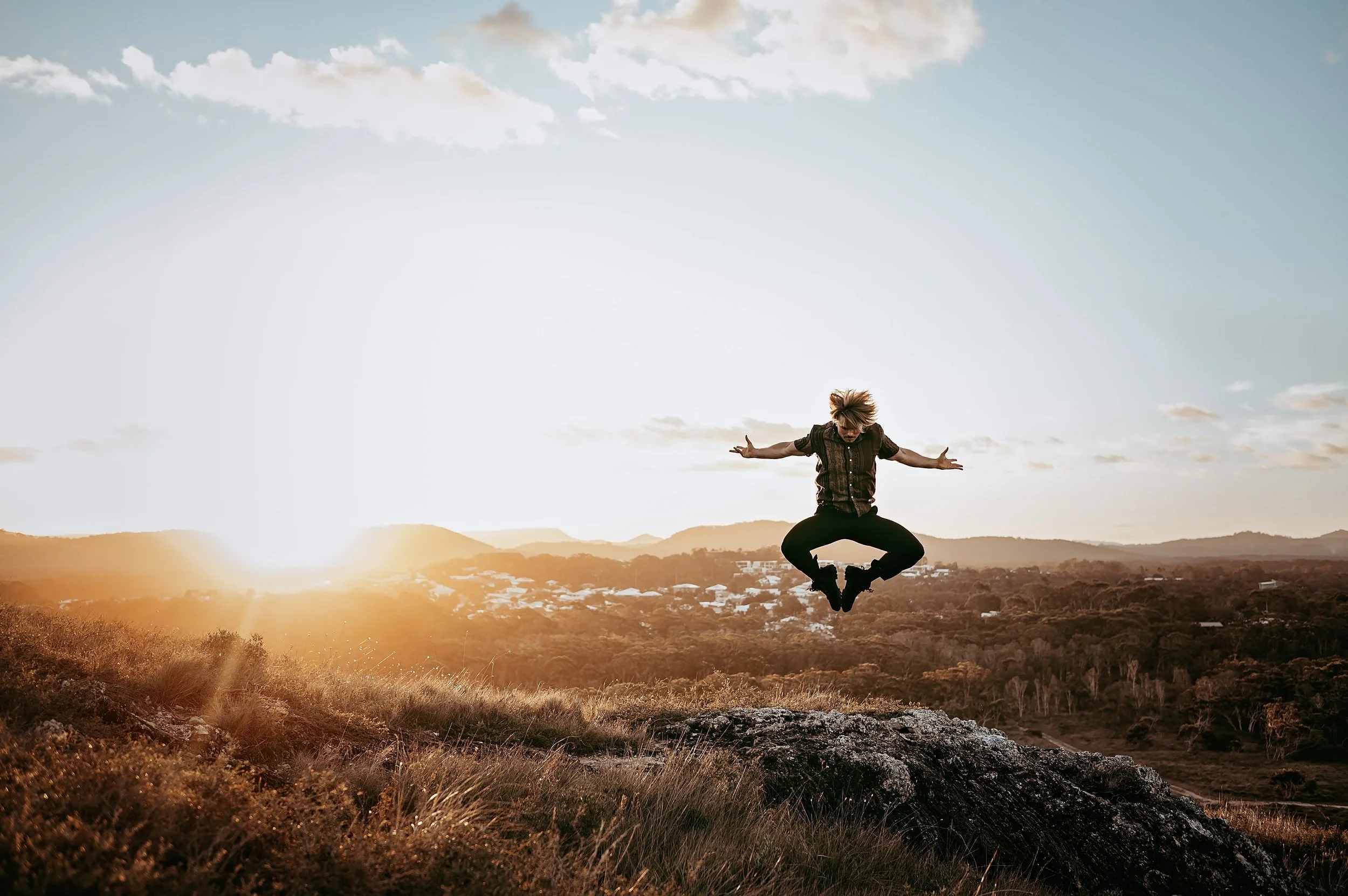 Jackson Kubian jumping in mid-air on a grassy hill during sunset with a scenic landscape and clouds in the sky.