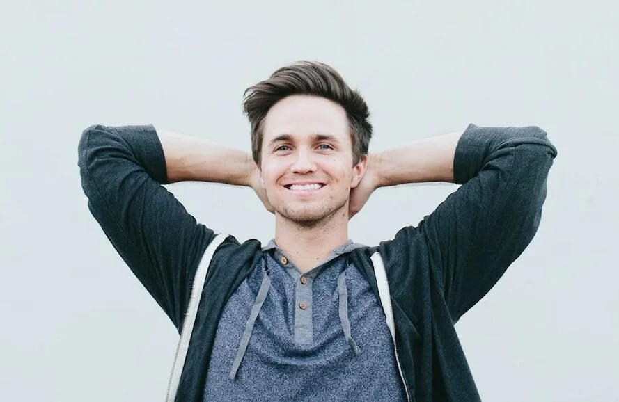 Smiling young man with short dark hair, wearing a dark hoodie over a blue shirt, standing against a plain light gray background.