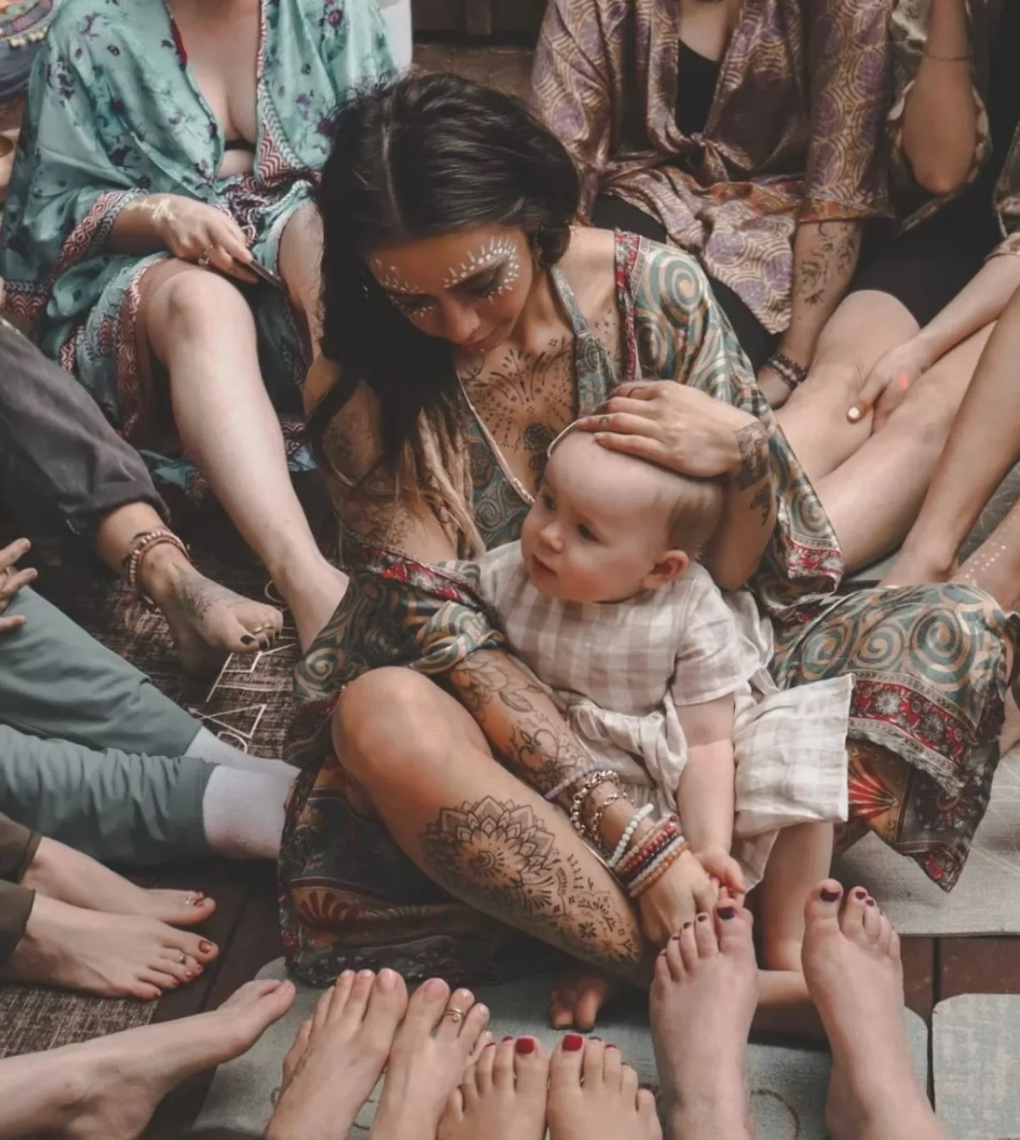 A woman with tattoos and face paint is sitting cross-legged, holding a young child on her lap during a gathering, surrounded by several barefoot people.