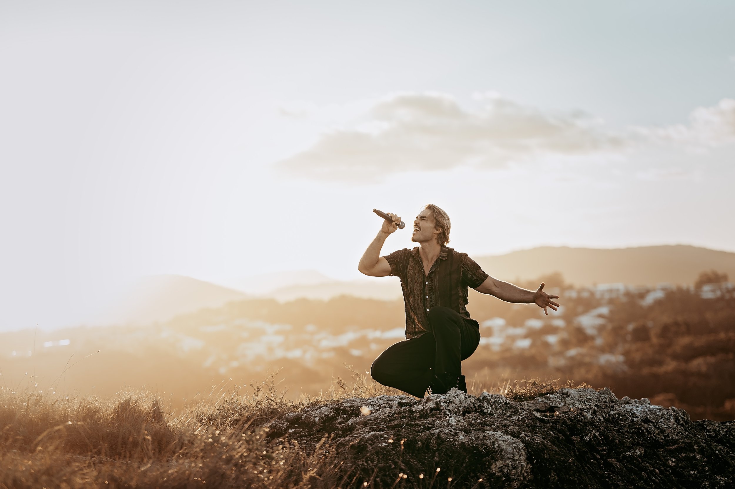 Jackson Kubian kneeling on a rock, singing into a microphone with a scenic outdoor background during sunset.