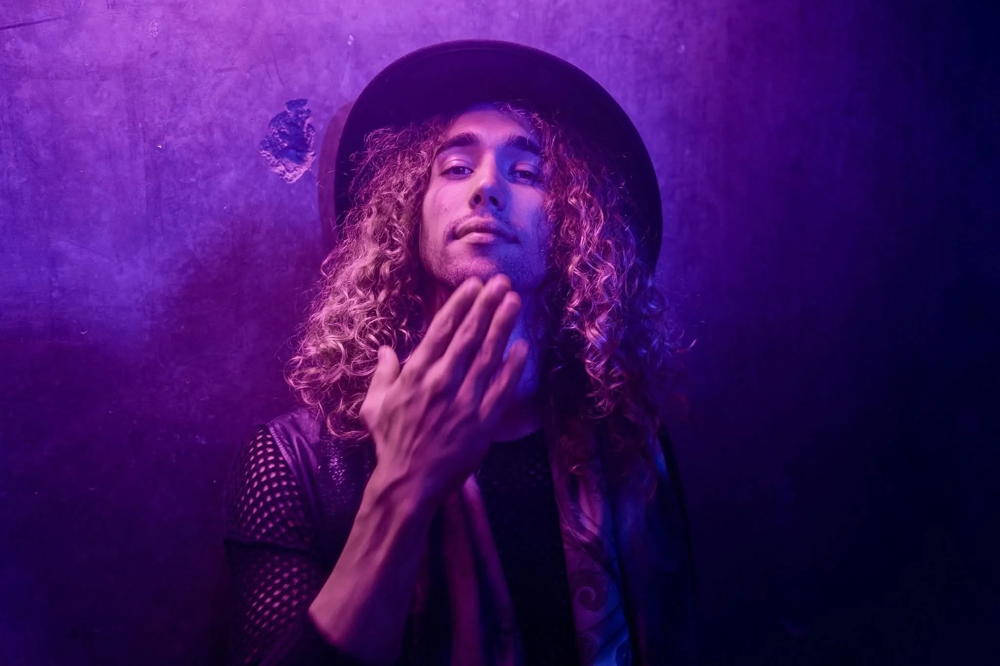 A person with long curly hair wearing a black hat, black mesh shirt, and a shiny vest, standing against a purple-lit background, touching their face with one hand and looking at the camera.