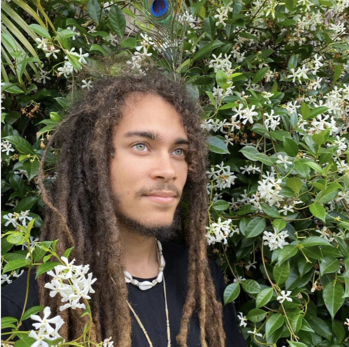 A young man with dreadlocks and light eyes standing in front of green foliage with white flowers and a peacock feather visible in the background.
