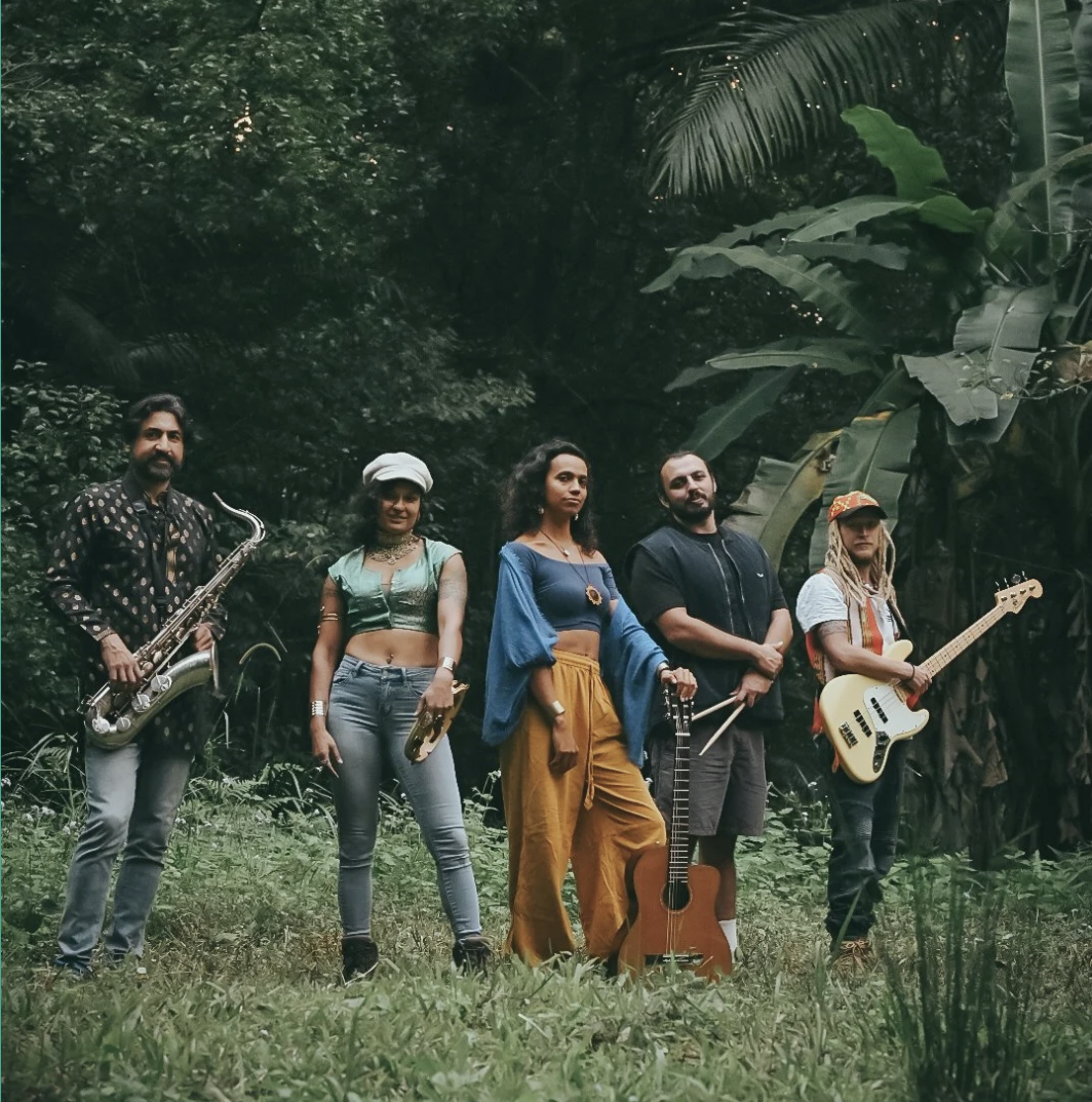 Solua a group of five musicians standing outdoors in a lush green environment, each holding a different musical instrument, with some looking at the camera and others looking away.