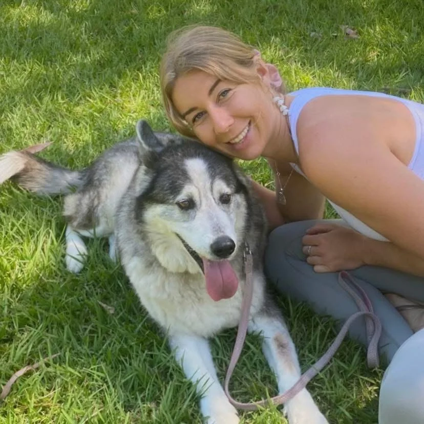A smiling woman lying on grass next to a Siberian Husky dog with a black and white coat.