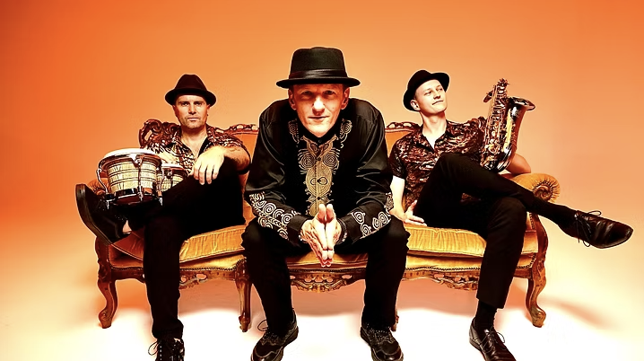 Three men sitting on an ornate vintage sofa against a gradient orange background. They are dressed in dark clothing with hats. The man in the middle is leaning forward with hands clasped, while the men on the sides hold musical instruments: a djembe drum on the left and a saxophone on the right.