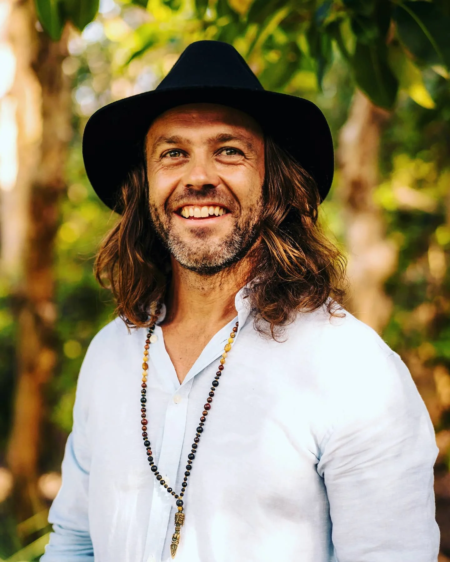 Smiling man with long hair, beard, and mustache, wearing a black wide-brimmed hat, white shirt, and beaded necklace, outdoors with green foliage in the background.