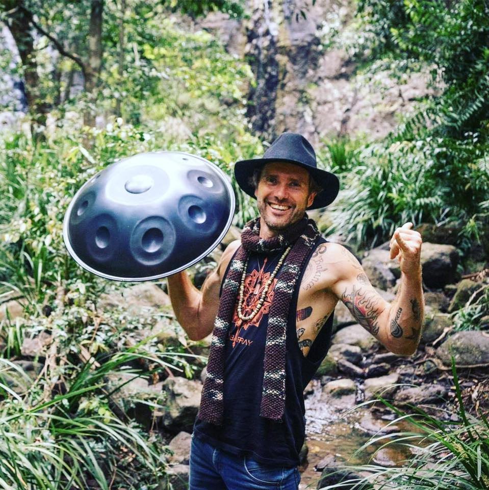 A man with tattoos, wearing a hat and sleeveless shirt, smiling and flexing his arm, holding a hang drum, stands in a lush green forest.