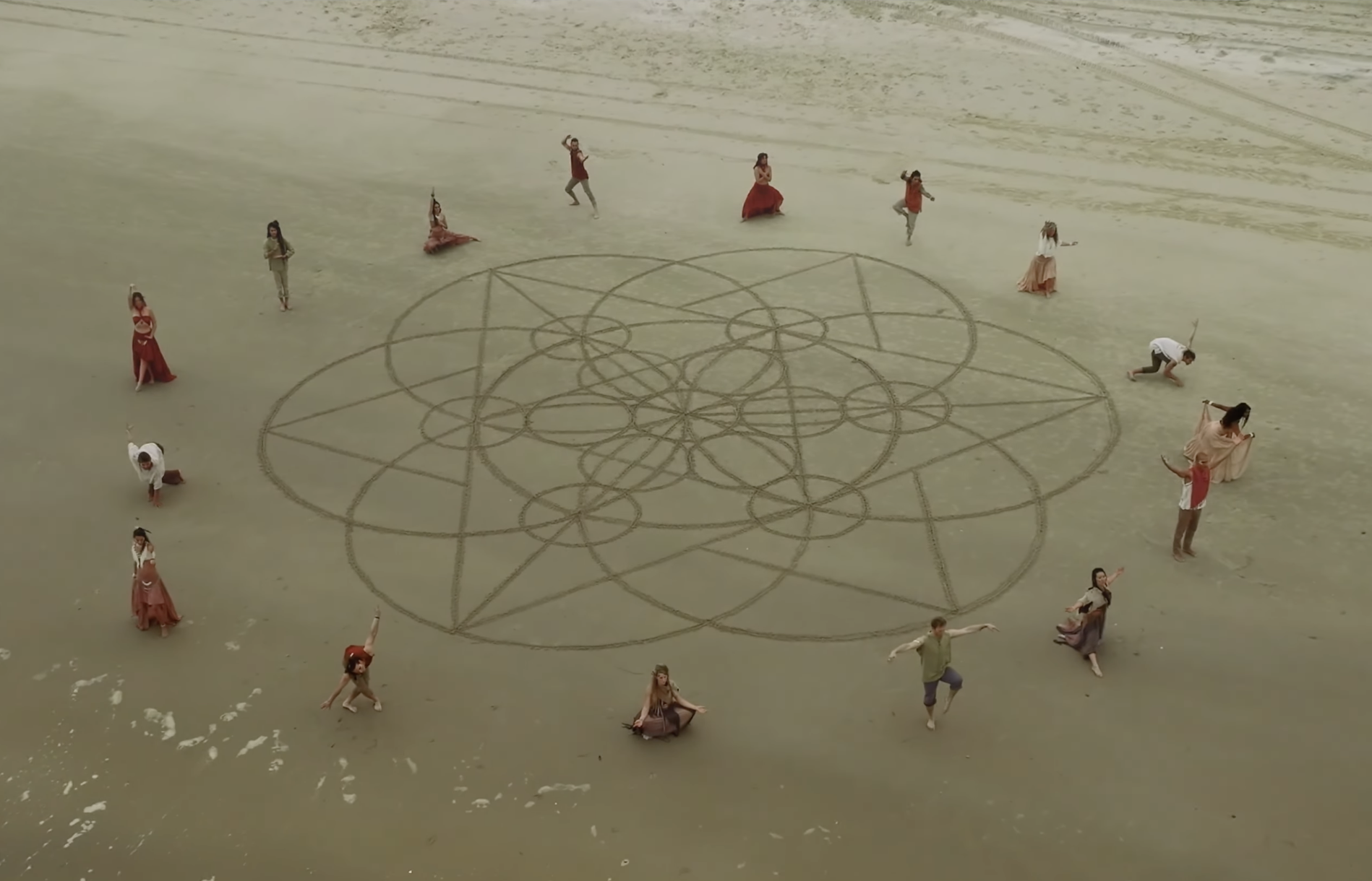 People standing and sitting on a beach around a large geometric sand art design, some posing with arms raised or extended.