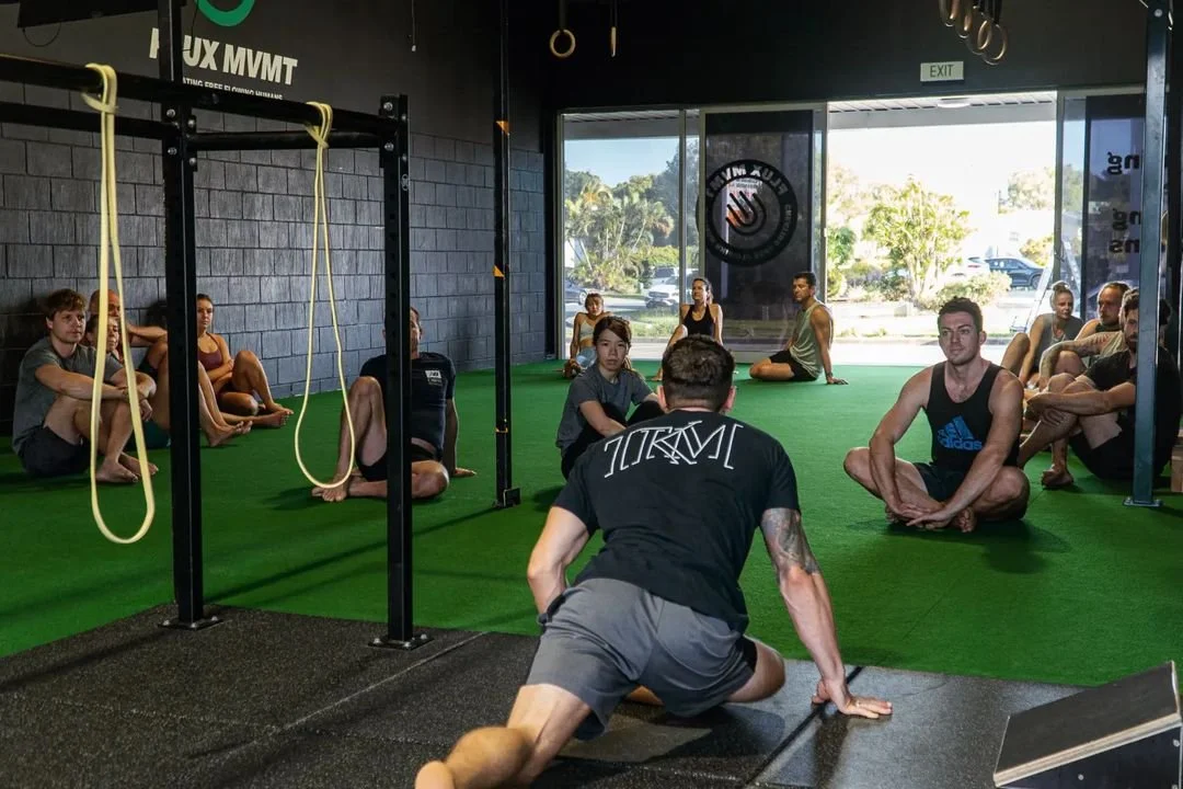 Kaizen Movement Group fitness class in gym with instructor leading seated participants on green artificial turf, gym equipment in background.