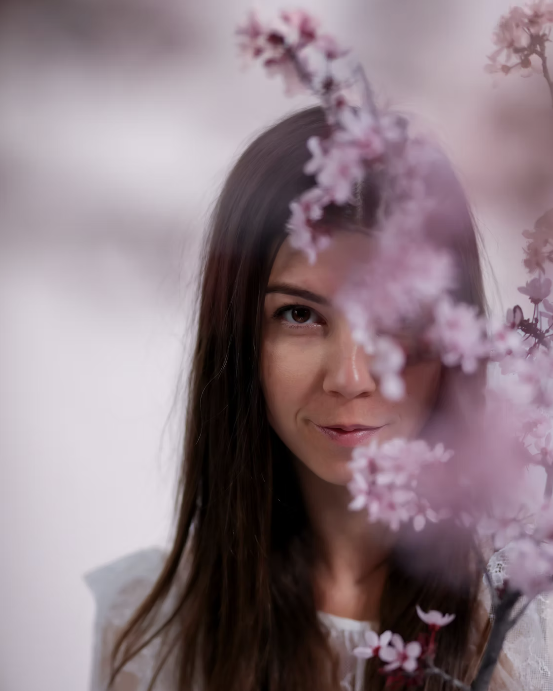 A woman with long brown hair partially obscured by pink cherry blossoms, smiling subtly at the camera.