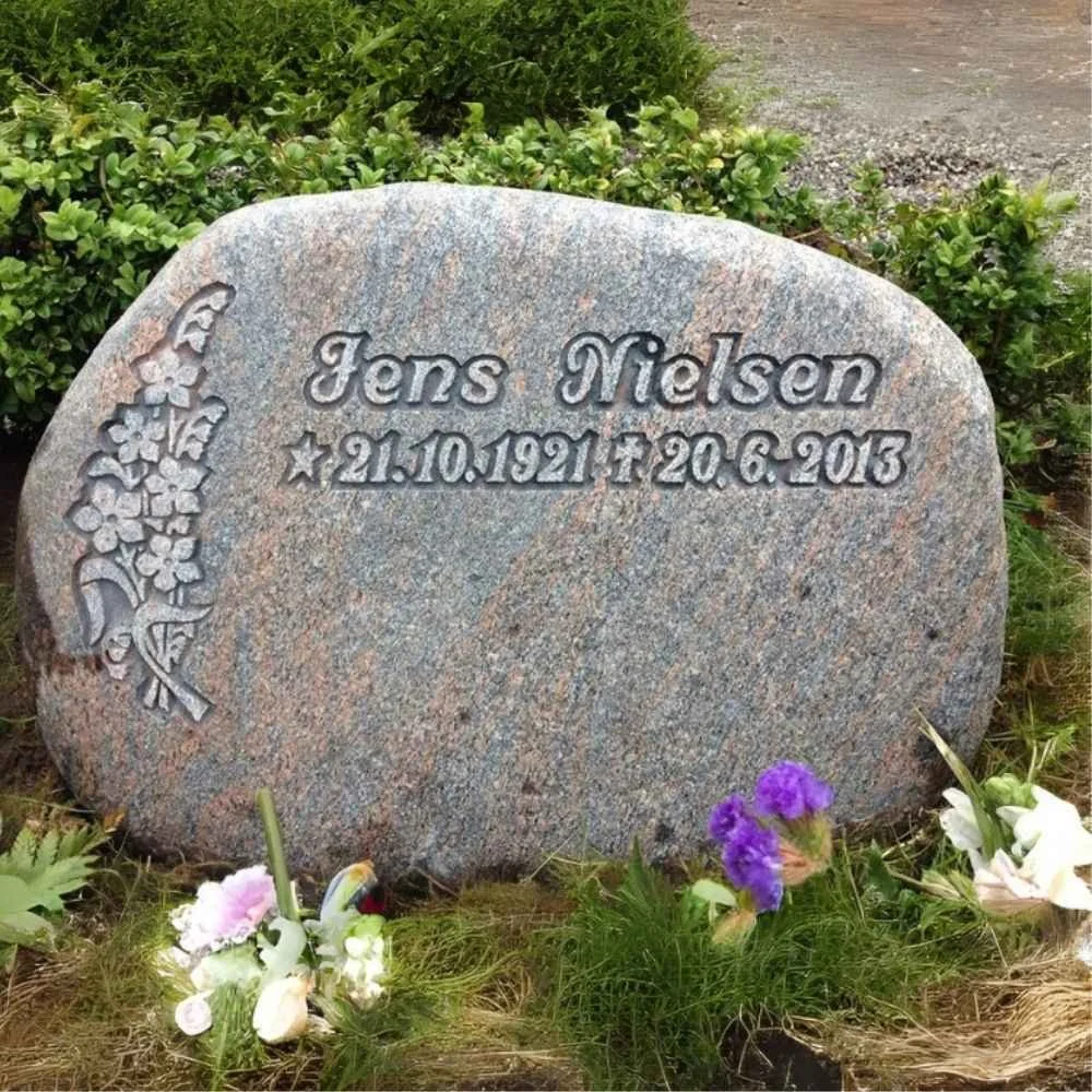Gravestone for Jens Nielsen with flower carvings on the left and surrounding purple and white flowers at the base.